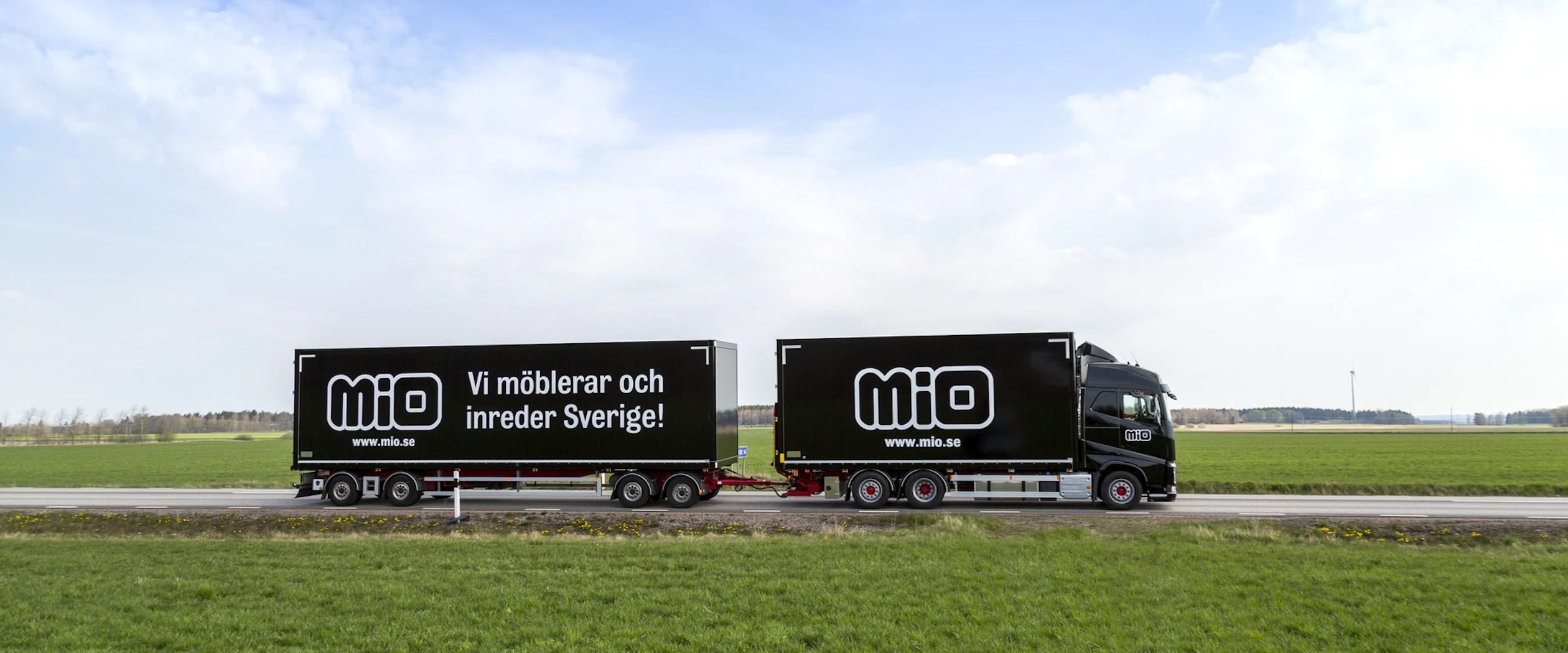 Svart lastbil med släp med Miologotyper på sidan och texten "Vi möblerar och inreder Sverige"
