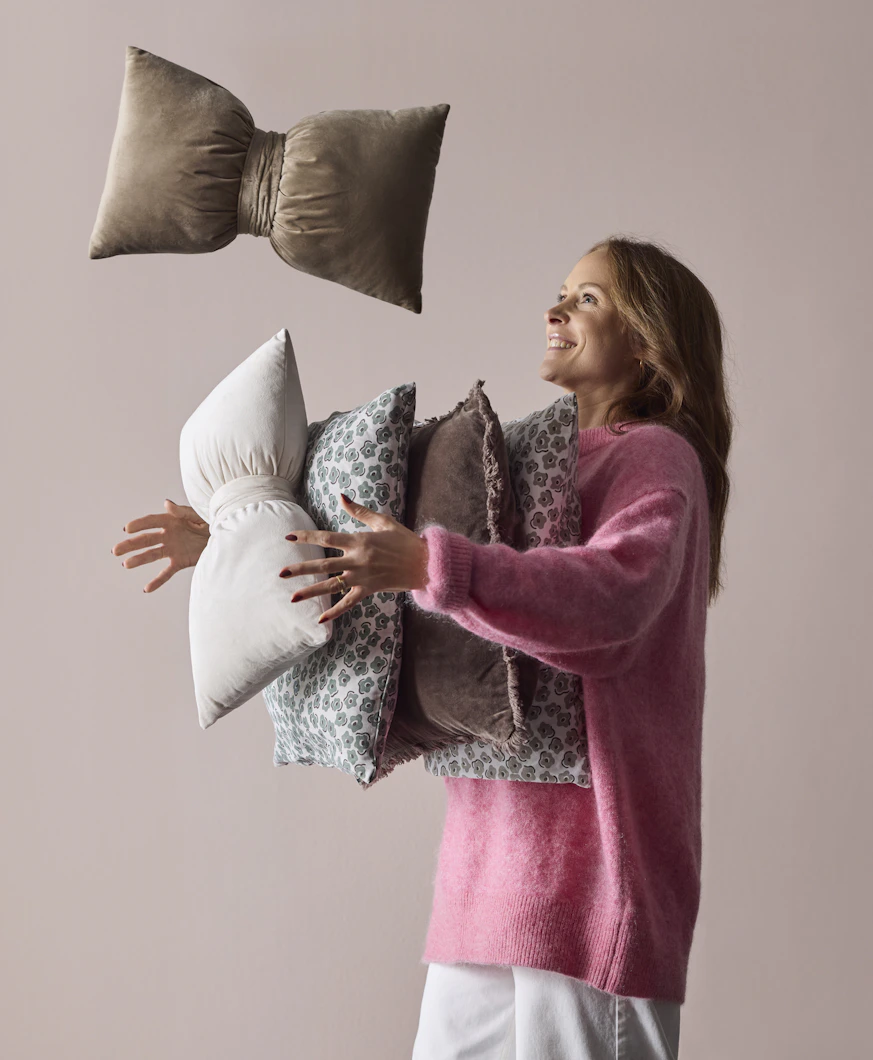 en tjej i rosa tröja står framför en rosa vägg och kastar upp prydnadskuddar i olika färger i luften