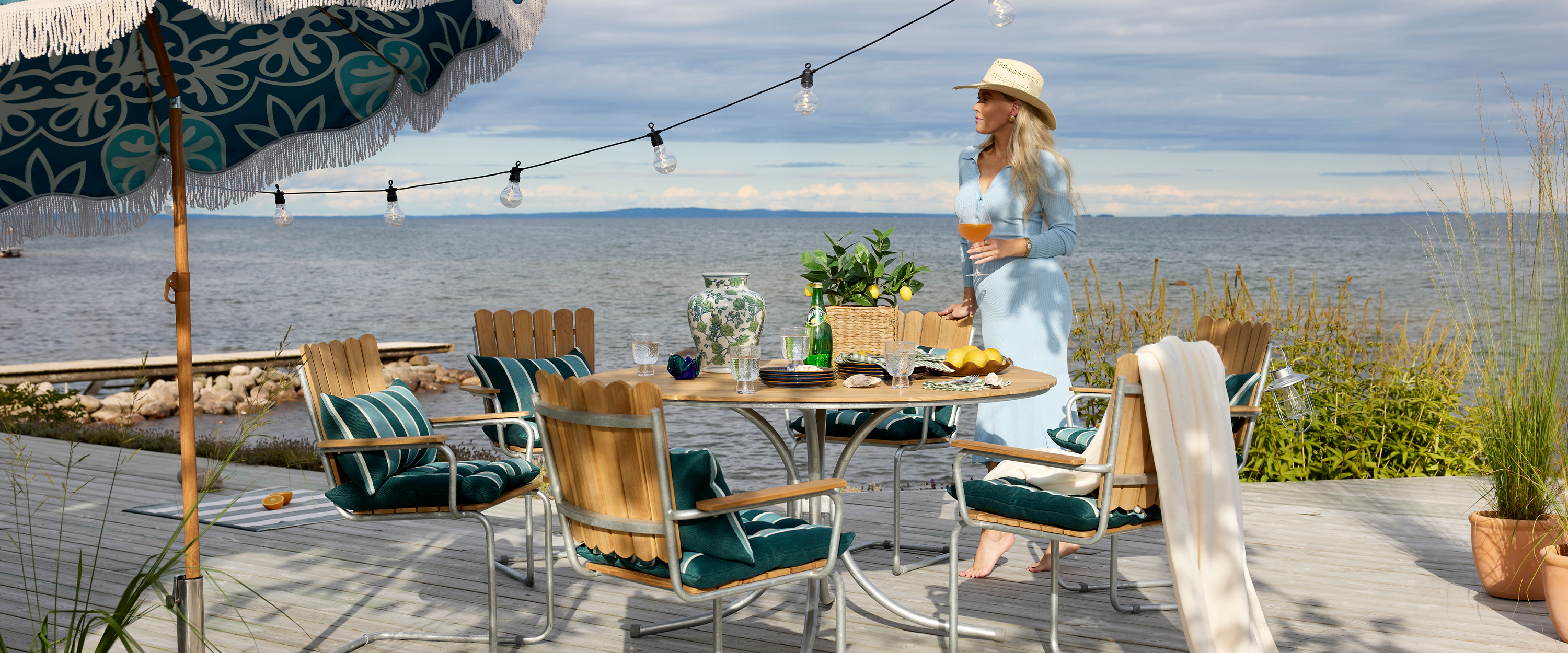 På en brygga står en kvinna intill en klassisk utegrupp i teak.