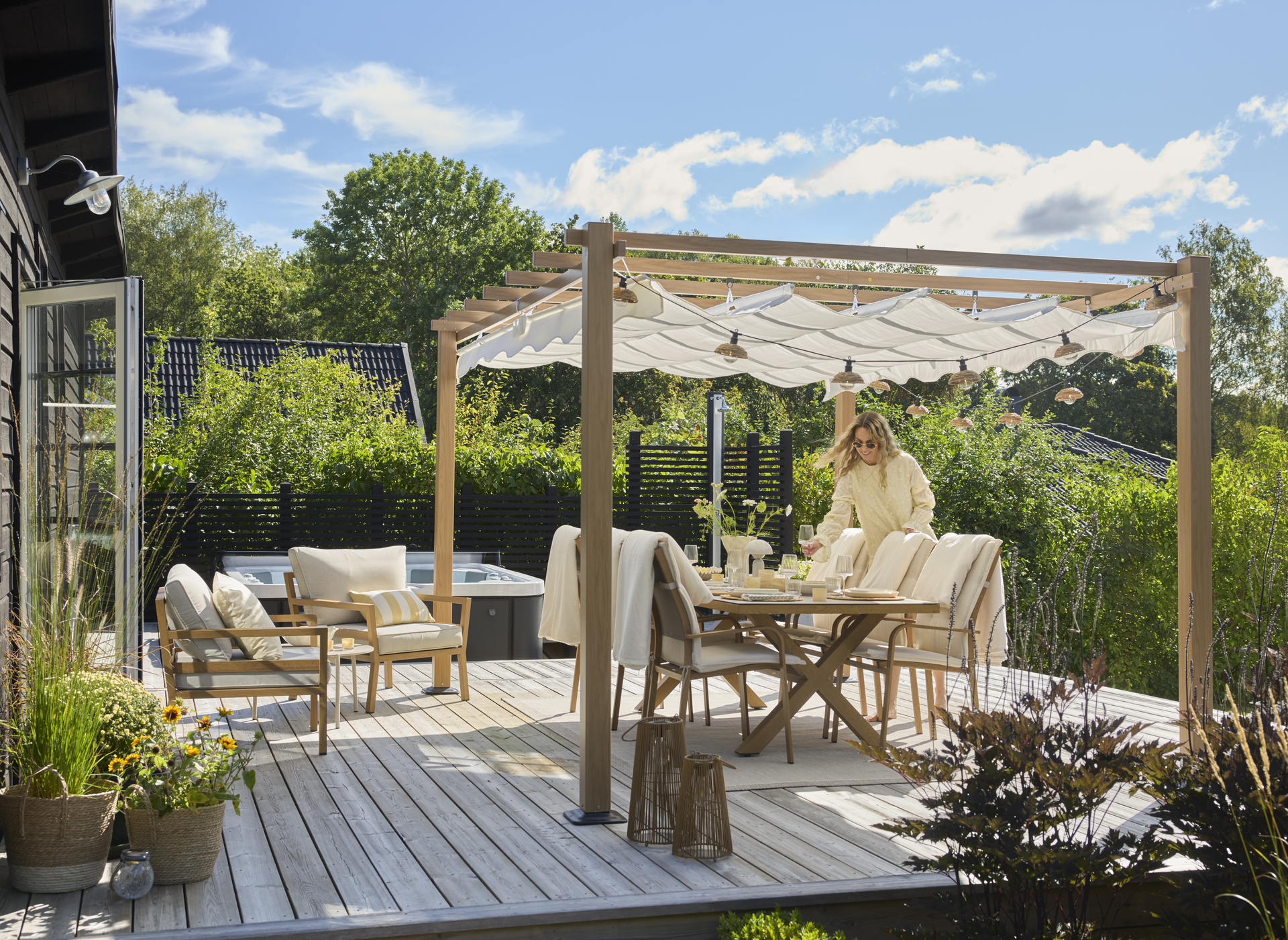 En kvinna dukar en utemöbelsgrupp som står på ett trädäck under en pergola.