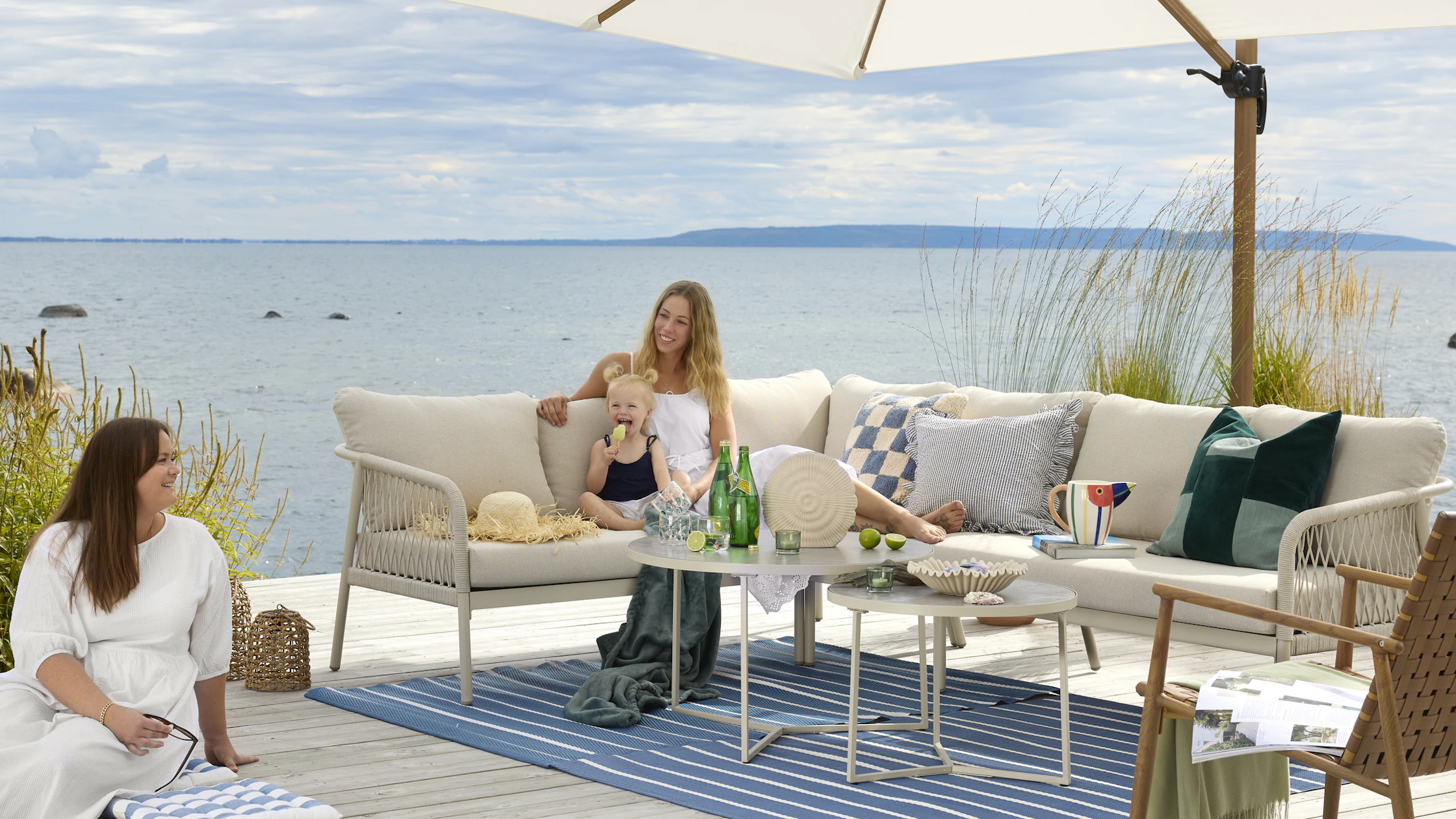 I en stor, beige utesoffa vid vattnet sitter två kvinnor och ett barn