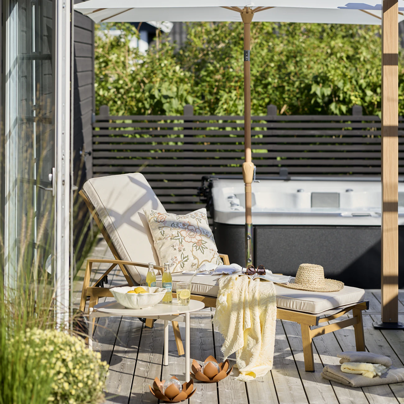 En stolstol med beiga dynor, blommig kudde och gul pläd står under ett parasoll på ett trädäck.