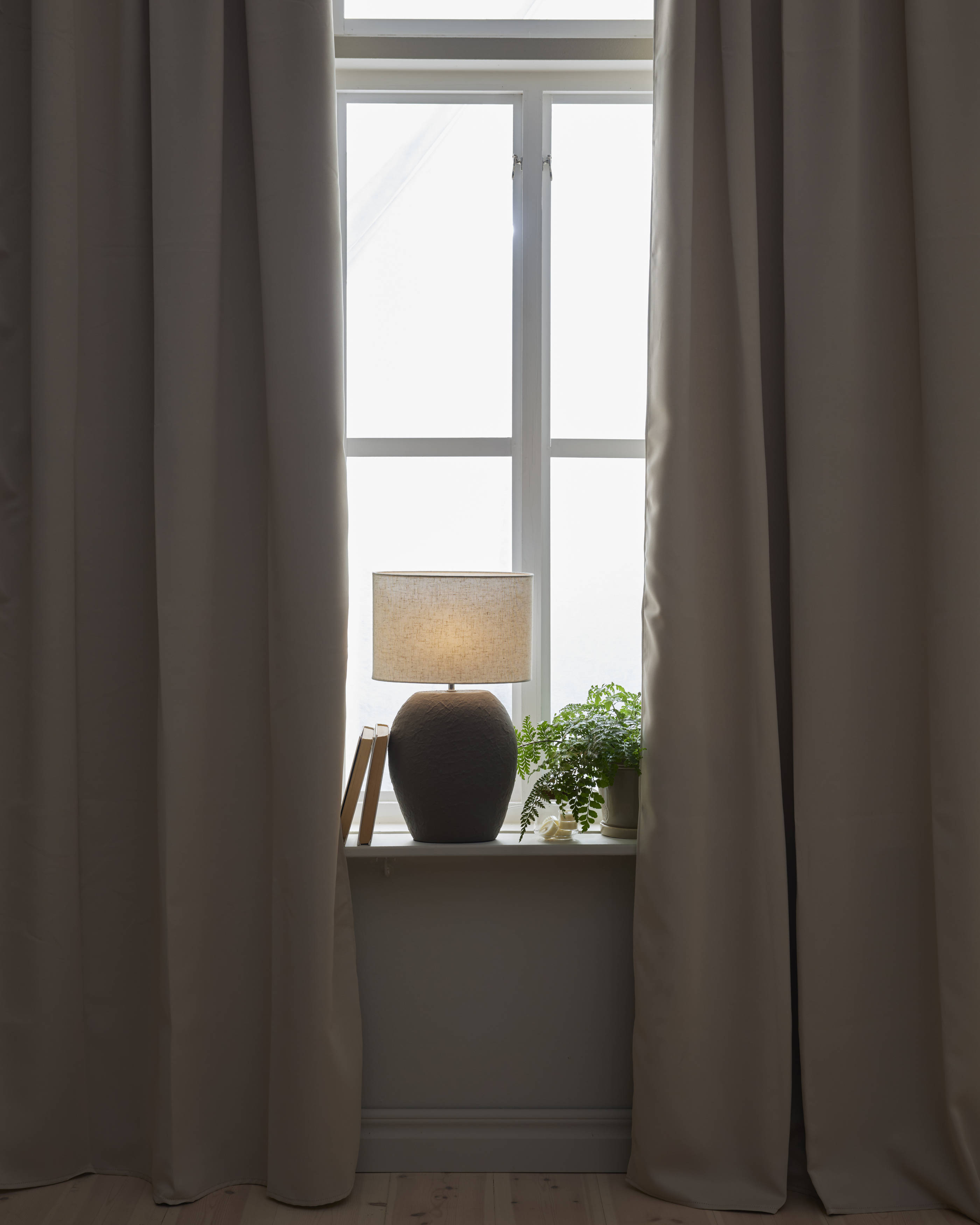 I ett fönster med mörkläggande, beiga gardiner står en mörkgrå lampfot med vit skärm