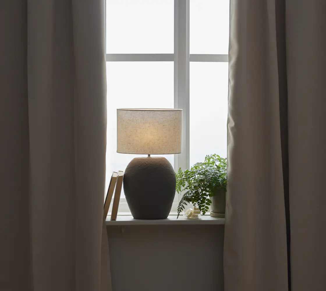 I ett fönster med mörkläggande, beiga gardiner står en mörkgrå lampfot med vit skärm