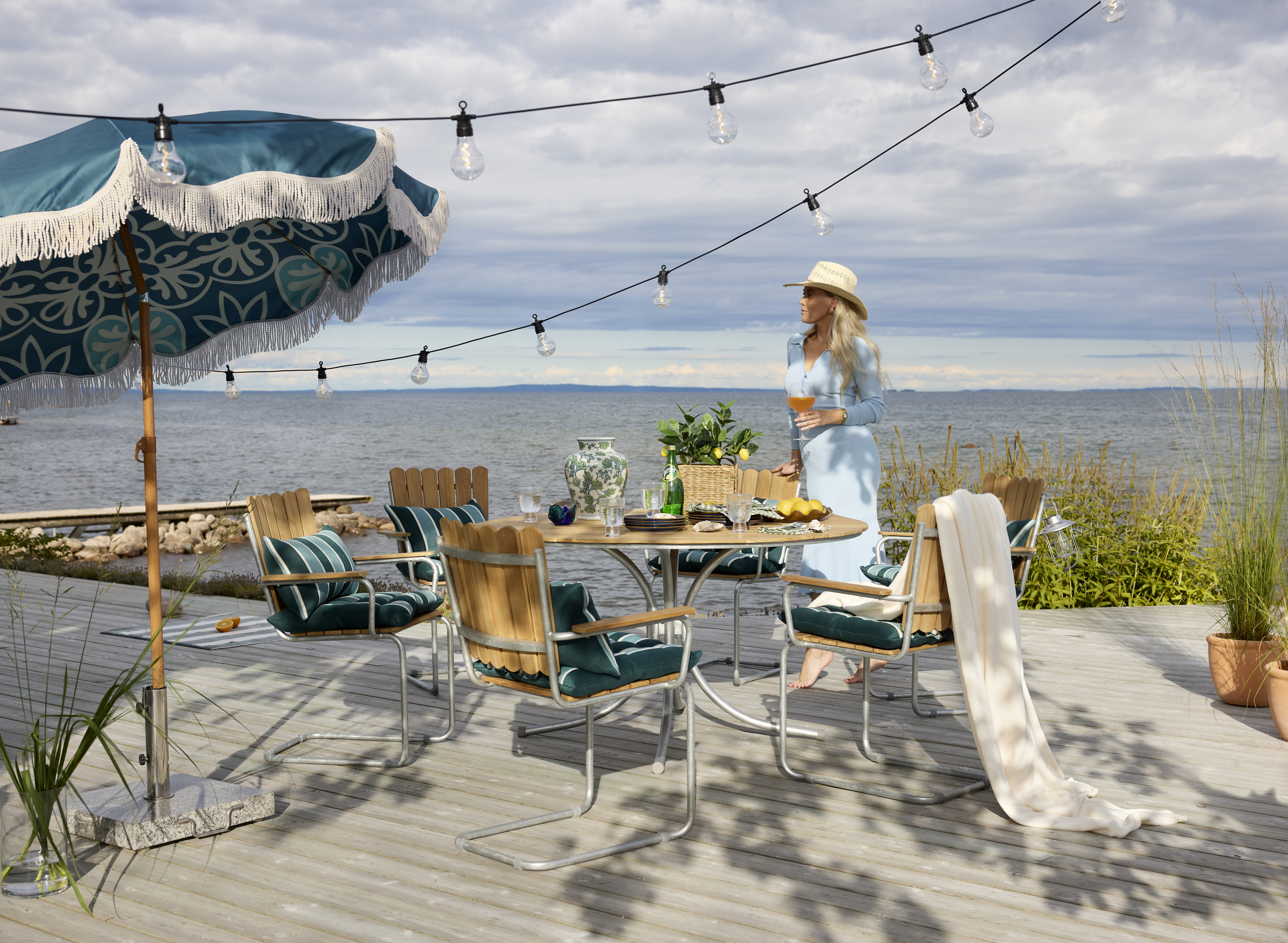 På en brygga står en kvinna intill en klassisk utegrupp i teak.