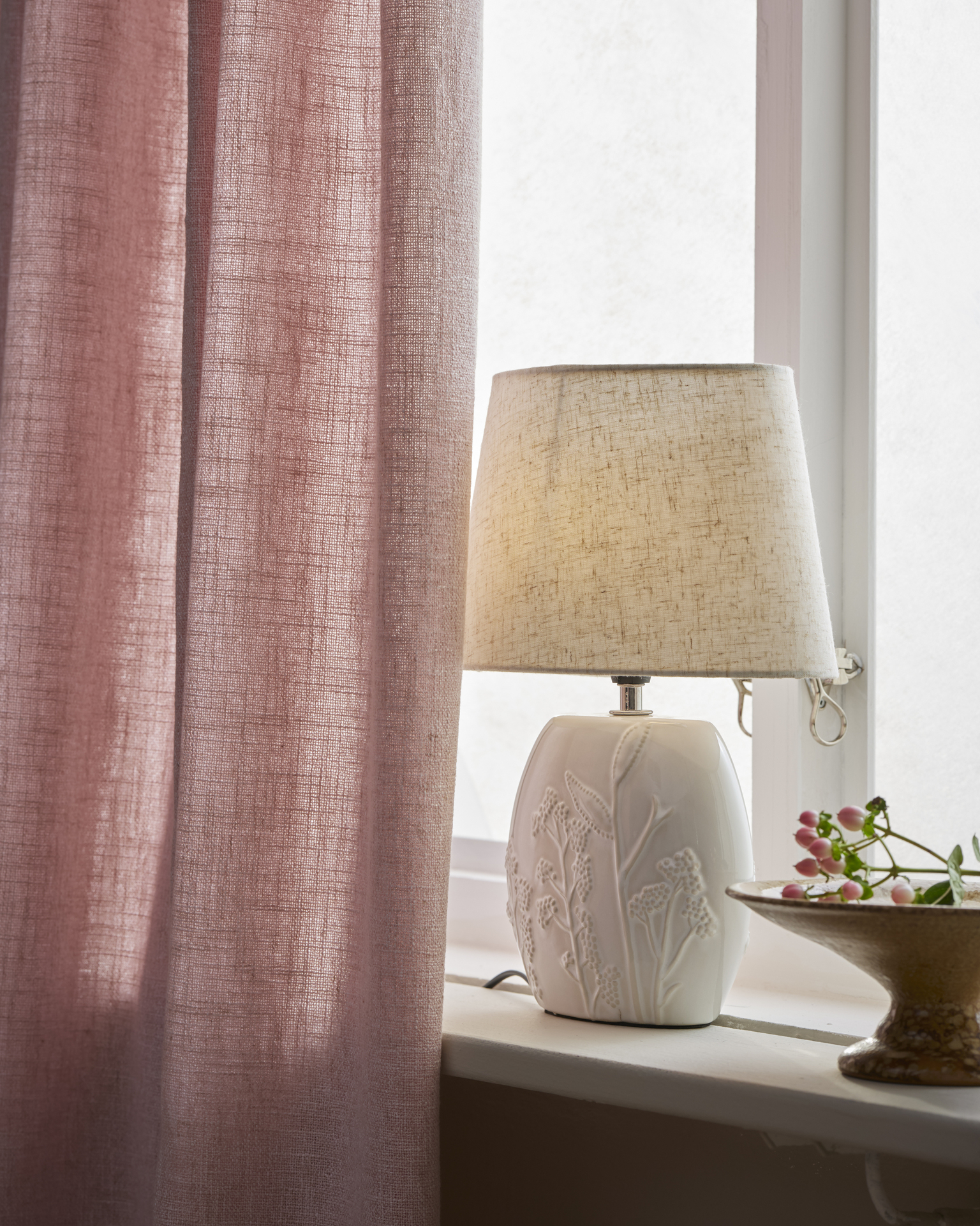 I ett fönster står en vit lampfot, till hälften dold av en rosa gardin