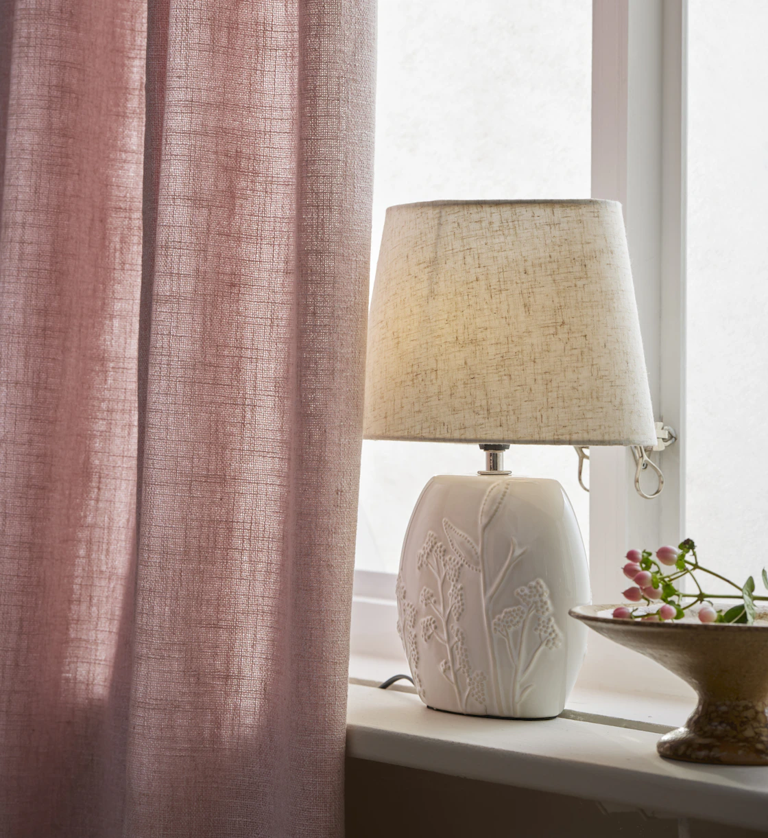 I ett fönster står en vit lampfot, till hälften dold av en rosa gardin