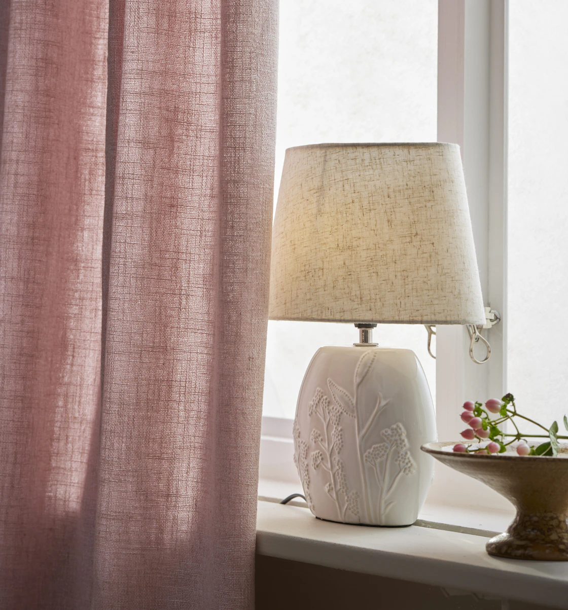 I ett fönster står en vit lampfot, till hälften dold av en rosa gardin