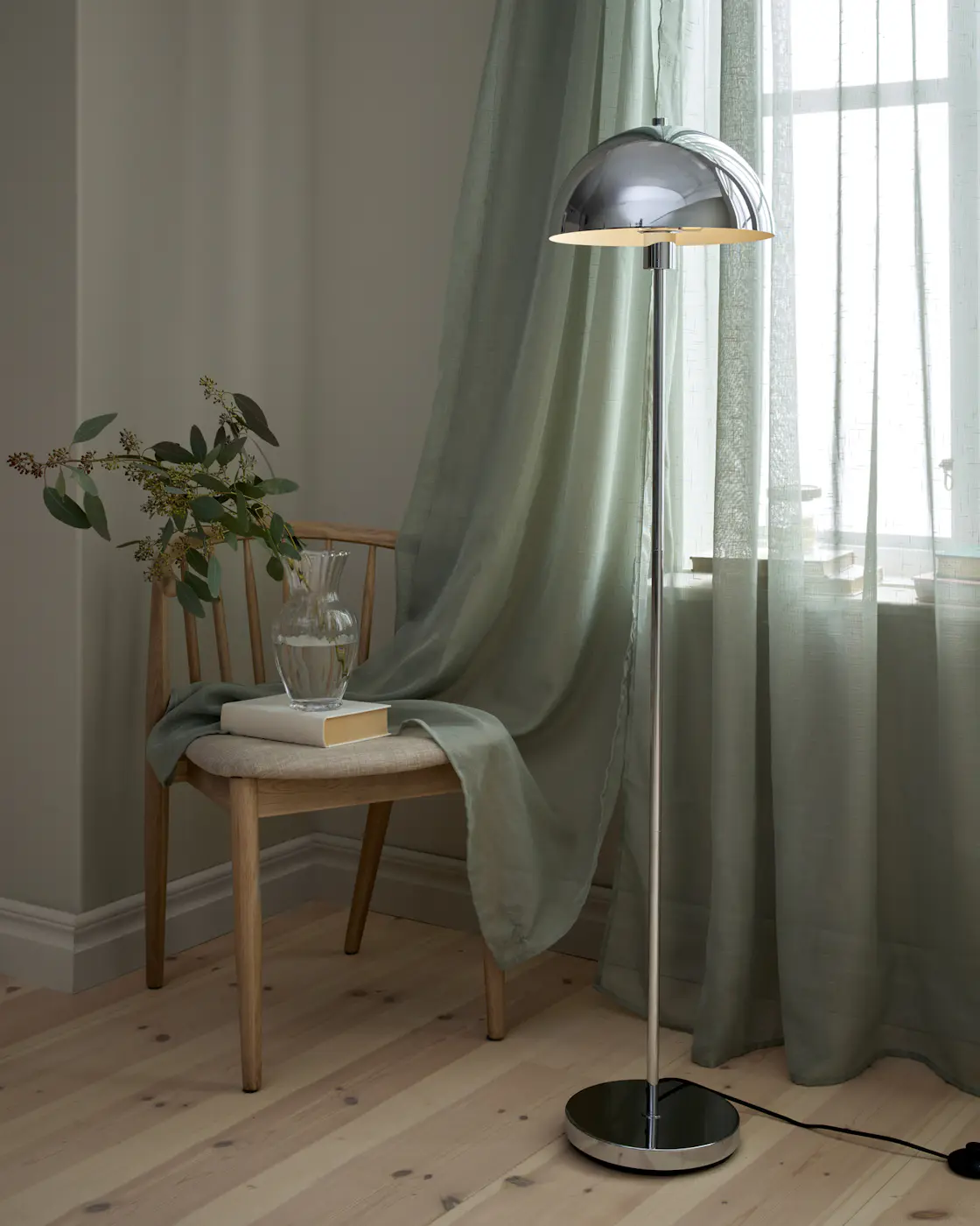 Framför ett fönster med skira, gröna gardiner står en golvlampa i chrome
