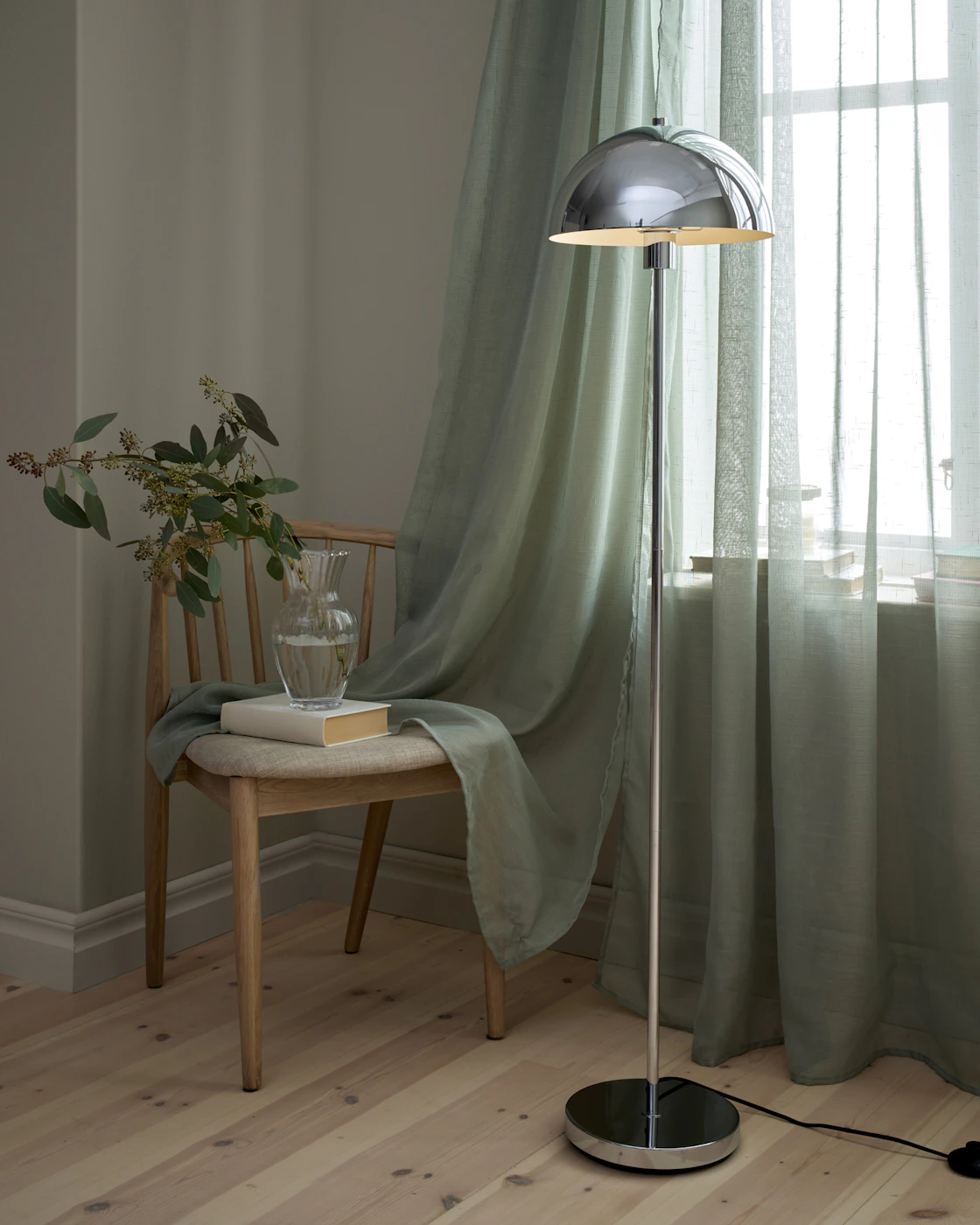 Framför ett fönster med skira, gröna gardiner står en golvlampa i chrome