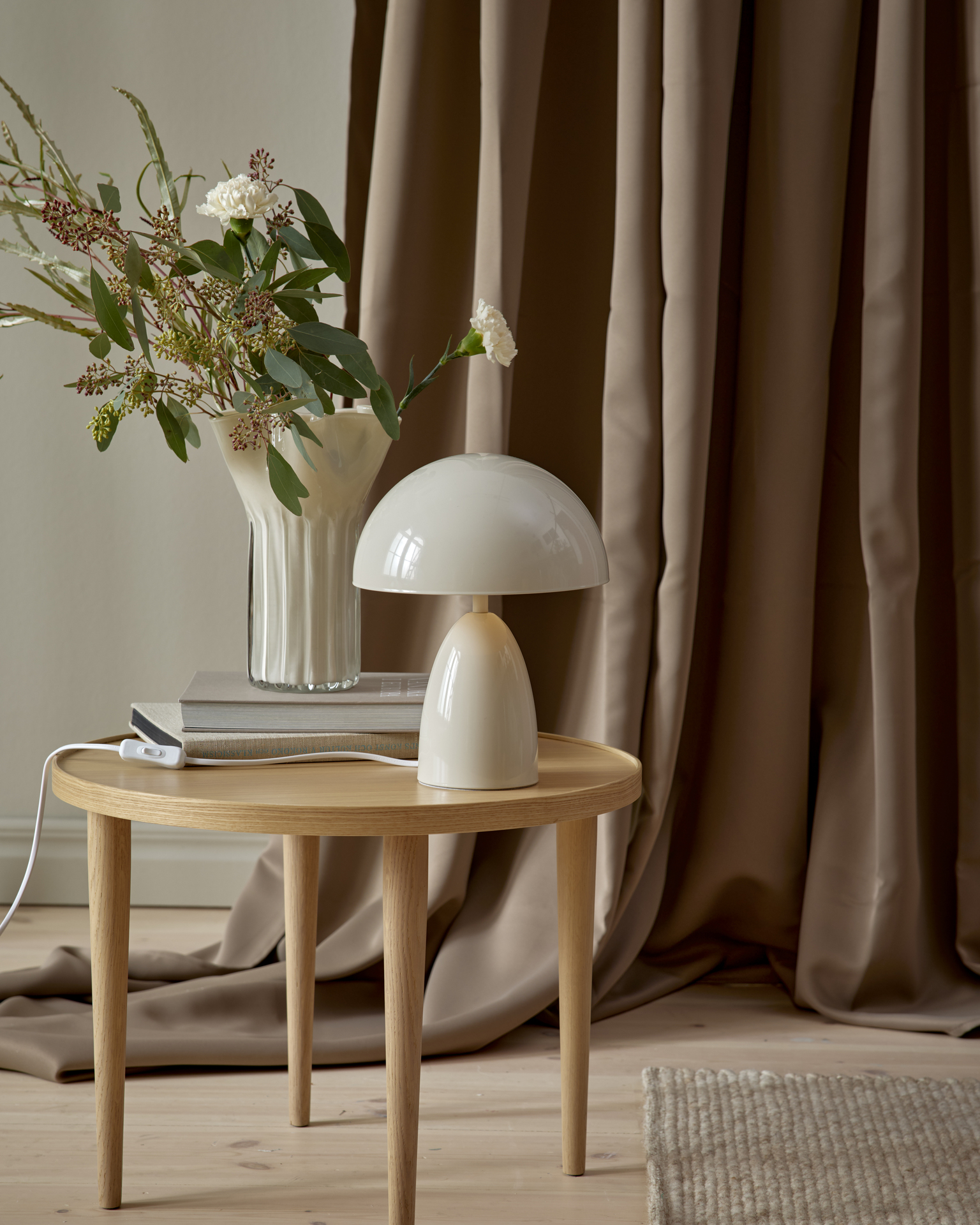 På ett litet sidobord i ljust trä står en modern, beige lampa. Bakom den är en brun gardin draperad