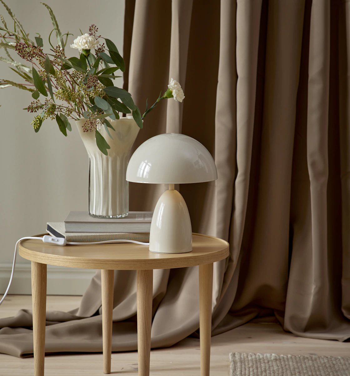 På ett litet sidobord i ljust trä står en modern, beige lampa. Bakom den är en brun gardin draperad