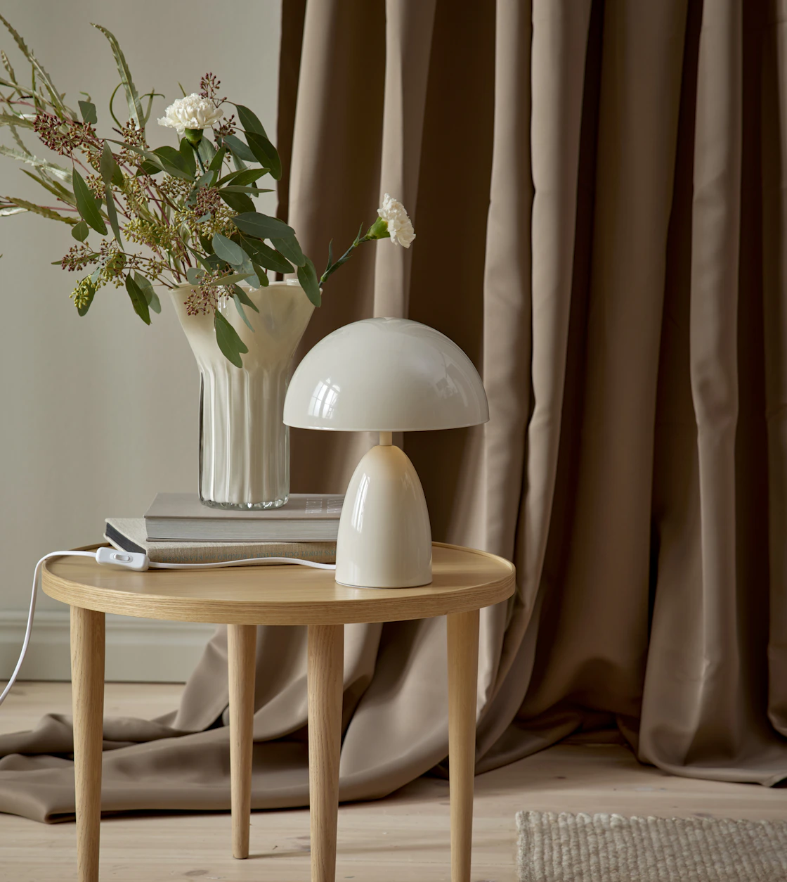 På ett litet sidobord i ljust trä står en modern, beige lampa. Bakom den är en brun gardin draperad
