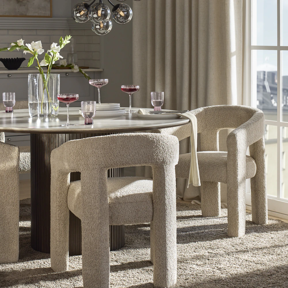 Ett runt bord i mörkt trä och fyra stolar i beige bouclé står i ett rum med ett stort fönster och ljusa gardiner.