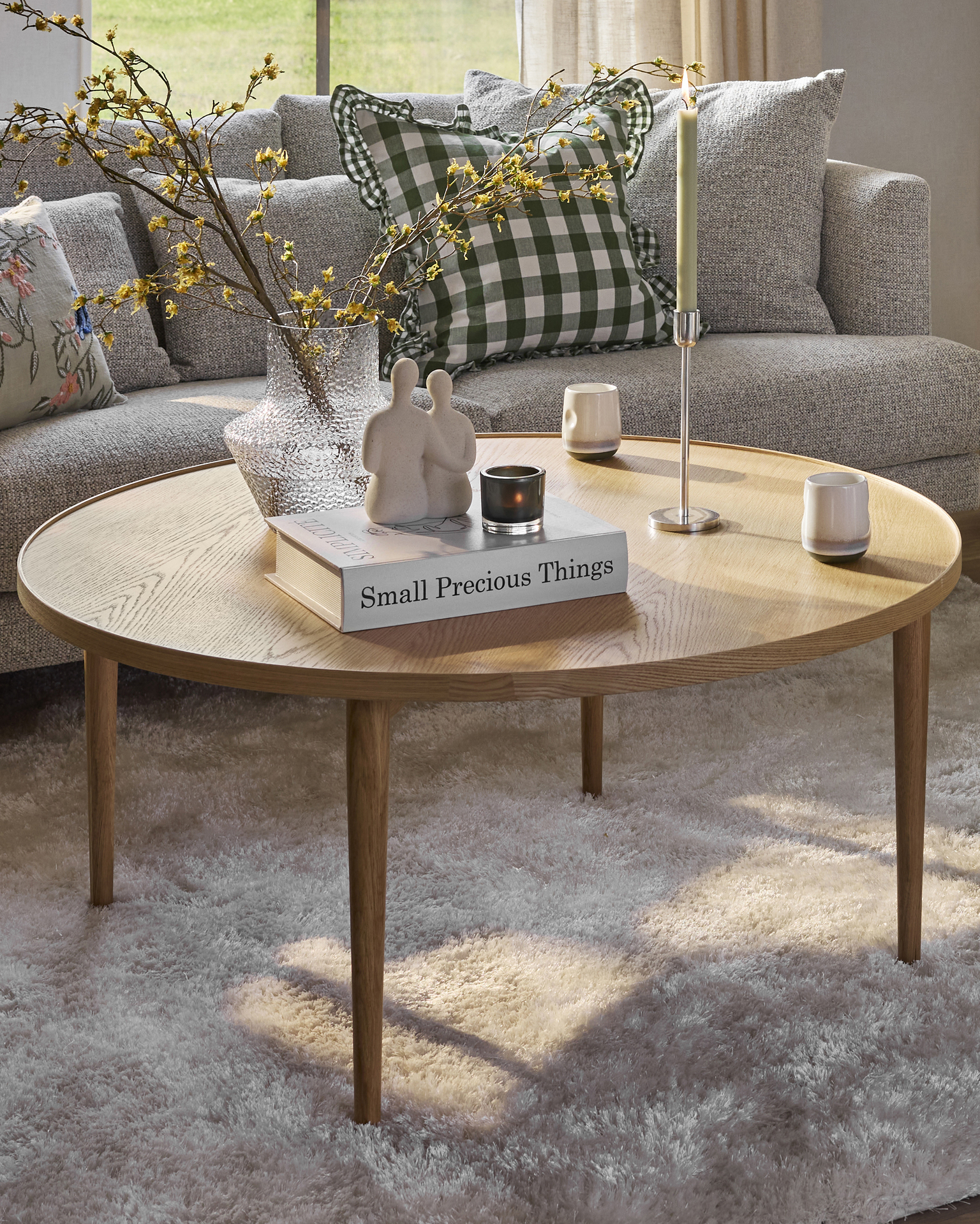 Ett runt ekfärgat soffbord står på en beige matta framför en grå soffa