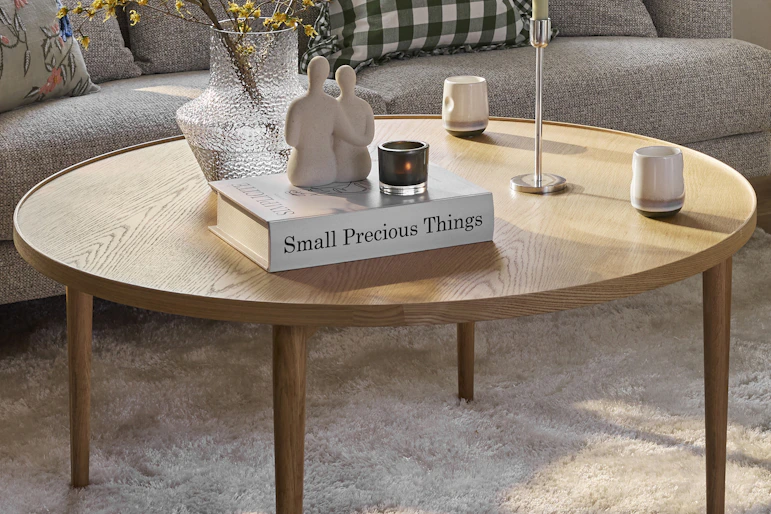 Ett runt ekfärgat soffbord står på en beige matta framför en grå soffa