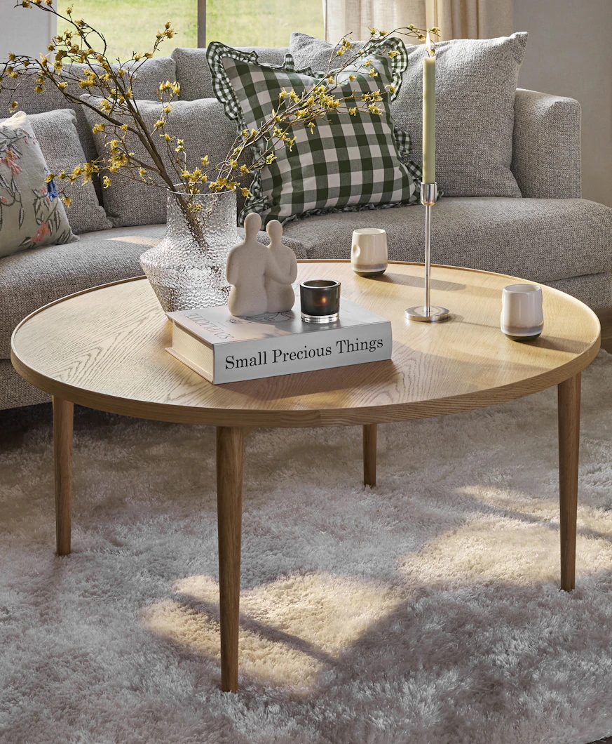 Ett runt ekfärgat soffbord står på en beige matta framför en grå soffa
