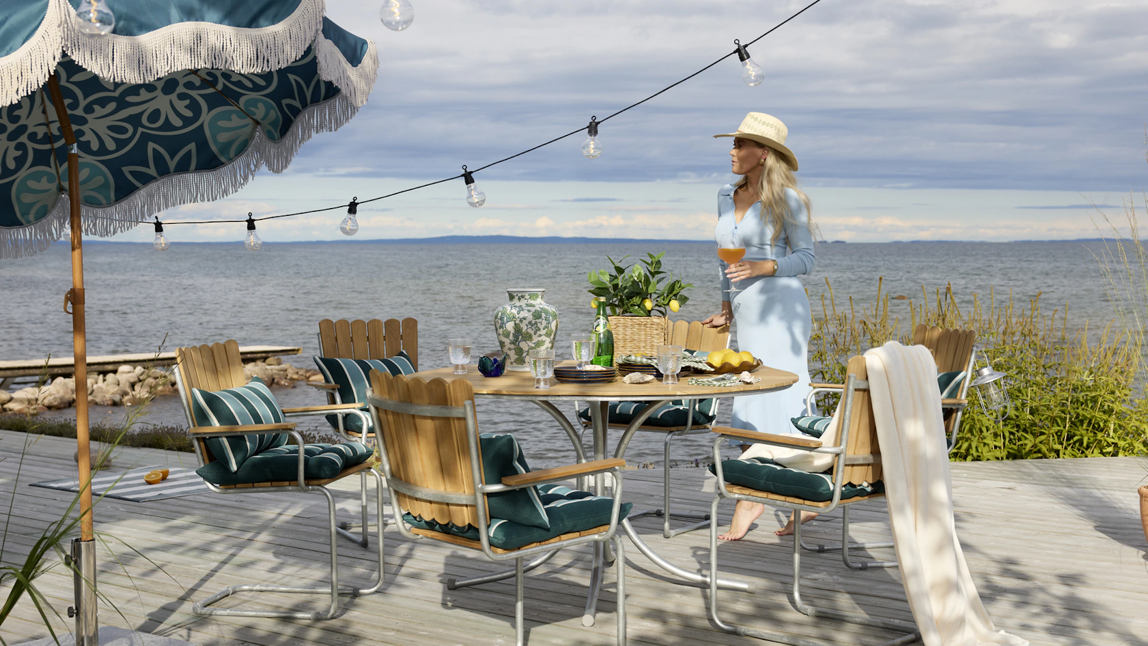 På en brygga står en kvinna intill en klassisk utegrupp i teak.