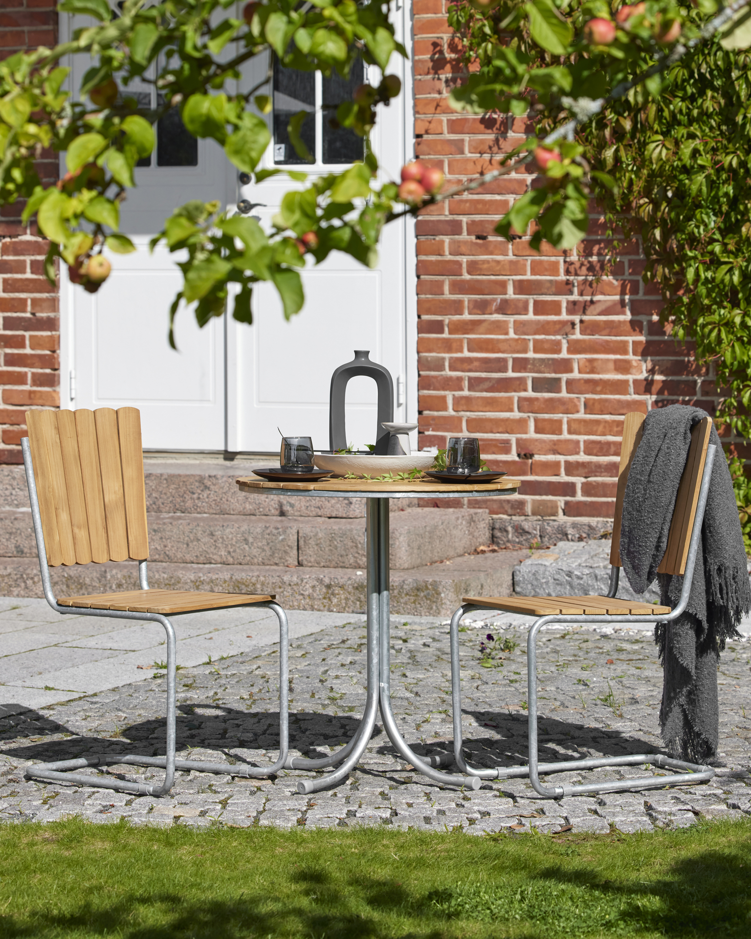 Ett caféset med bord och två stolar i teak står under ett träd.