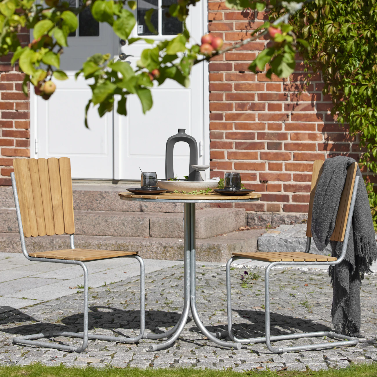 Ett caféset med bord och två stolar i teak står under ett träd.