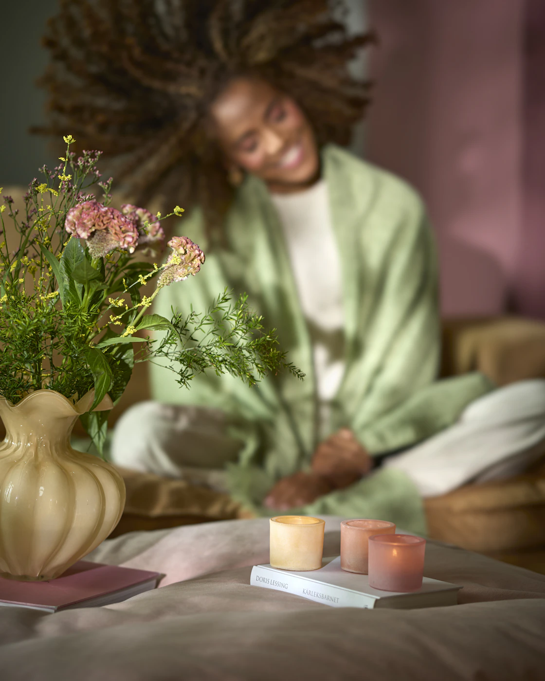 En gul vas och tre ljuslyktor är placerade på två böcker på en sittpuff. I bakgrunden sitter en kvinna.