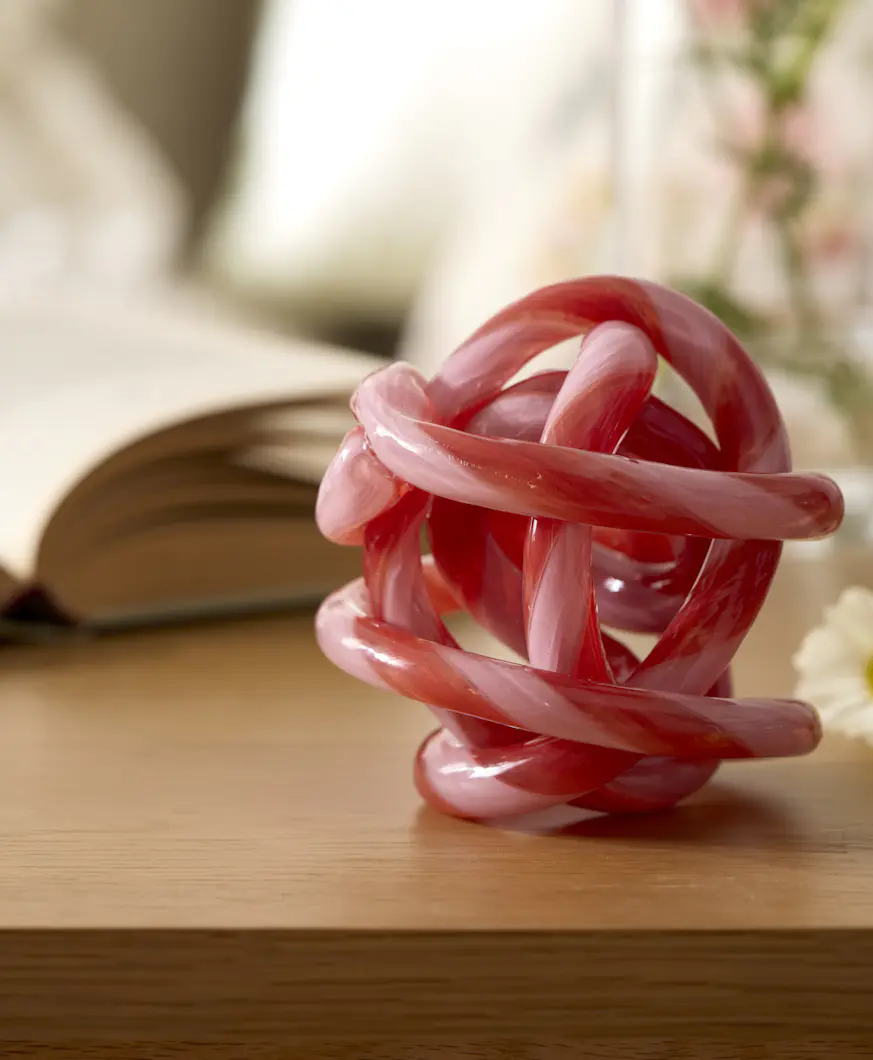 Rosa prydnadsföremål formad som en knut i glas står på ett bord framför en bok och en vas.