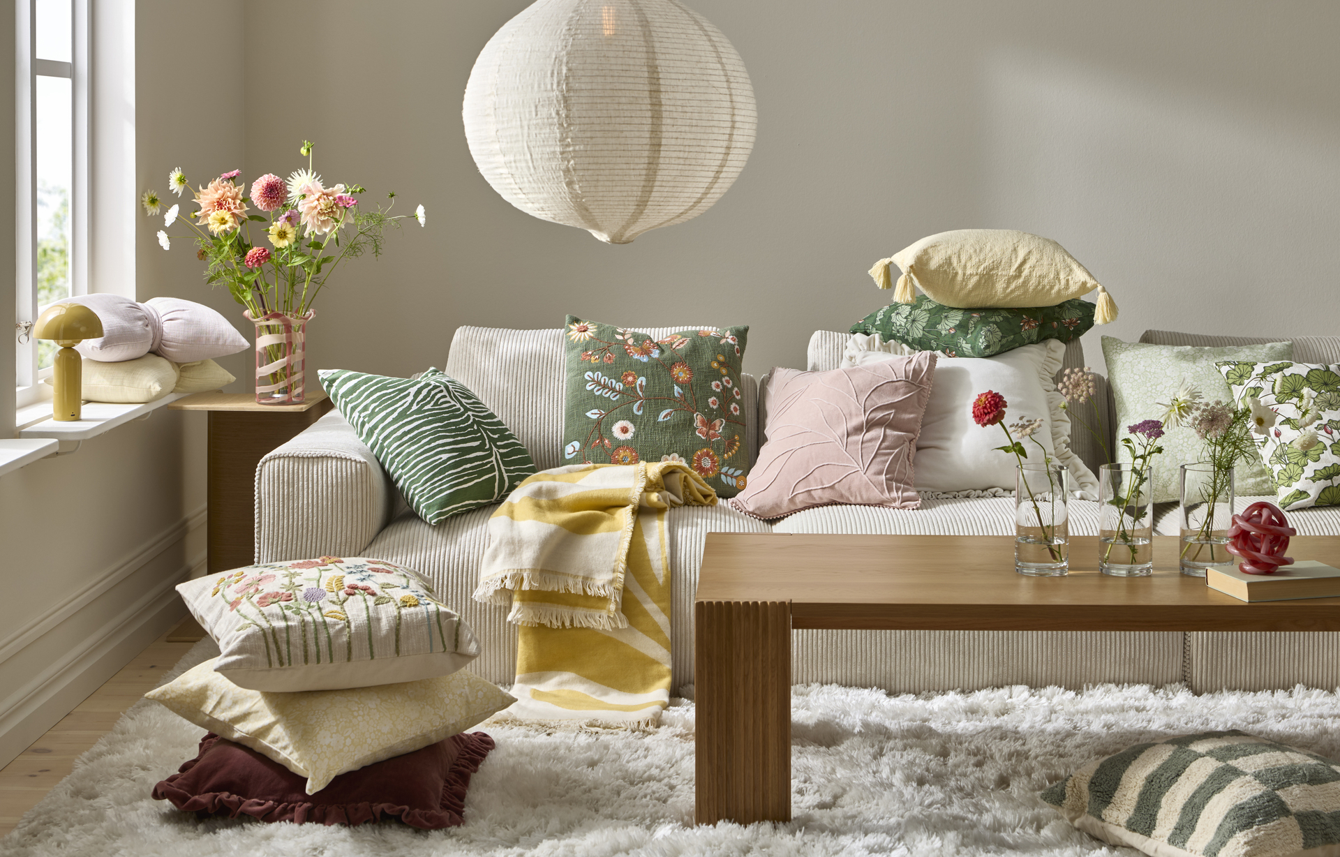 En ljus soffa full av kuddar och plädar i färgglada mönster. Till vänster om soffan finns ett bord med en färgglad bukett i en vas.