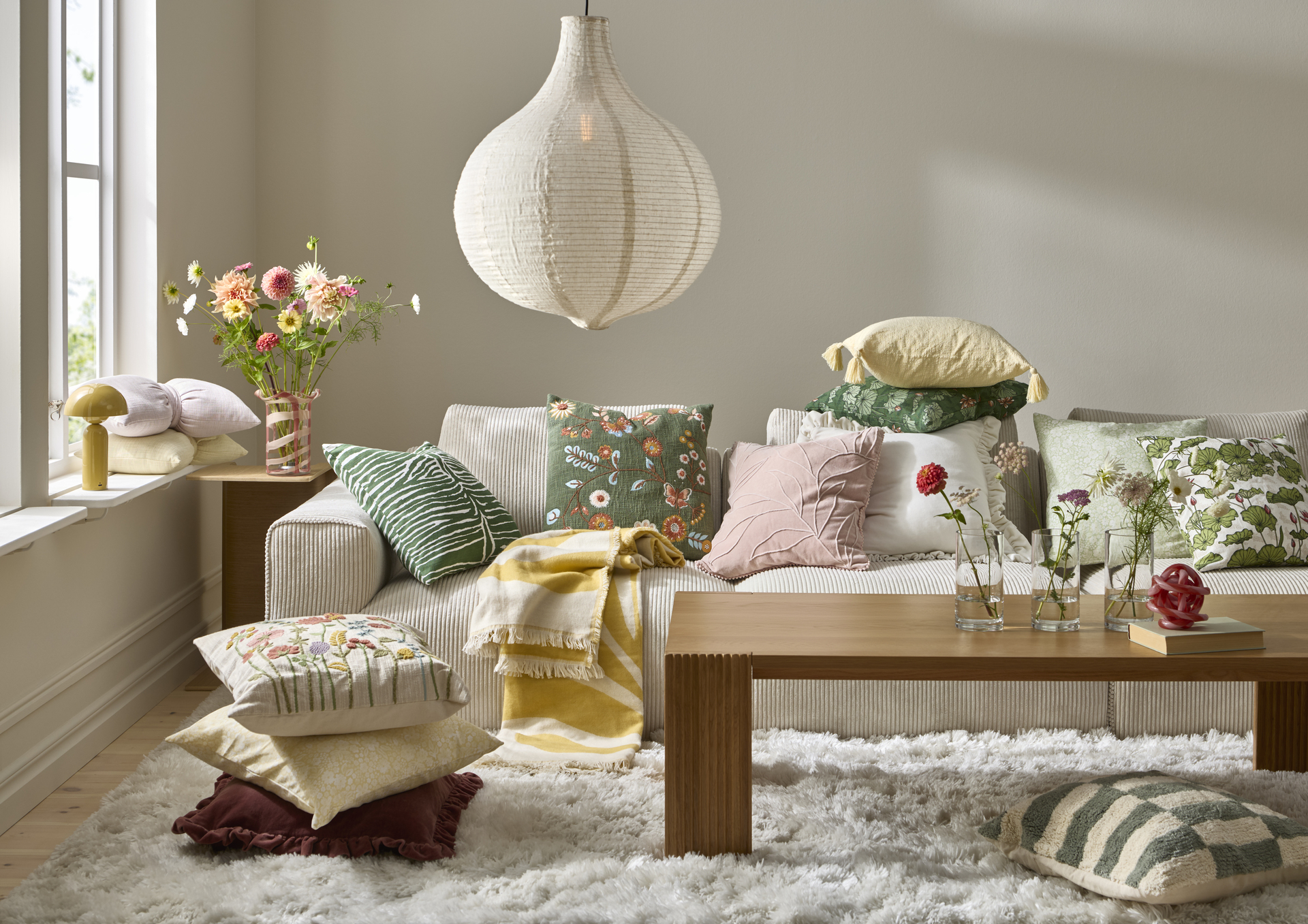 En ljus soffa full av kuddar och plädar i färgglada mönster. Till vänster om soffan finns ett bord med en färgglad bukett i en vas.