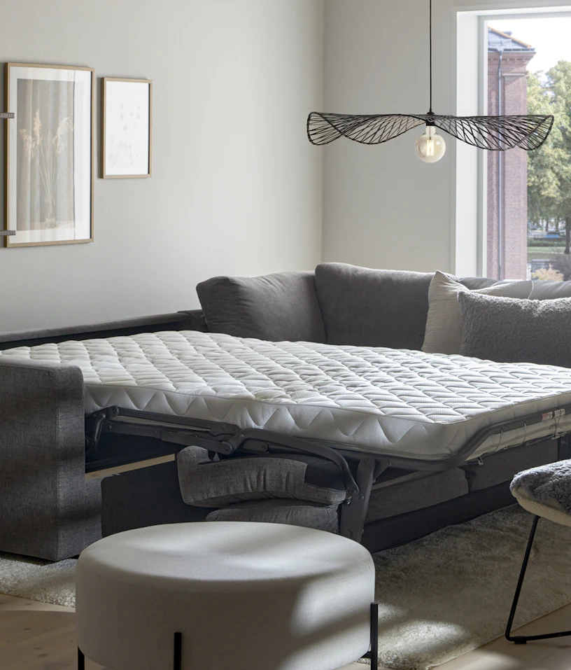 I ett vitt rum står en grå bäddsoffa med madrass utbäddad