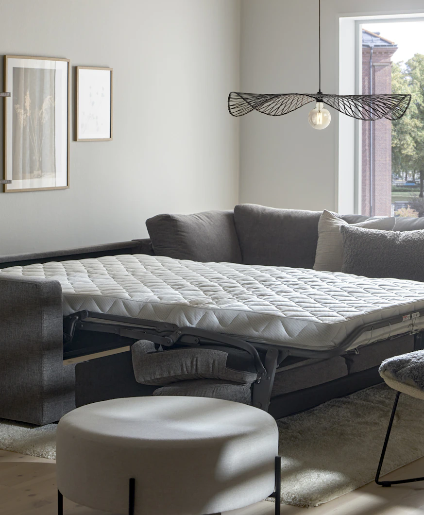 I ett vitt rum står en grå bäddsoffa med madrass utbäddad