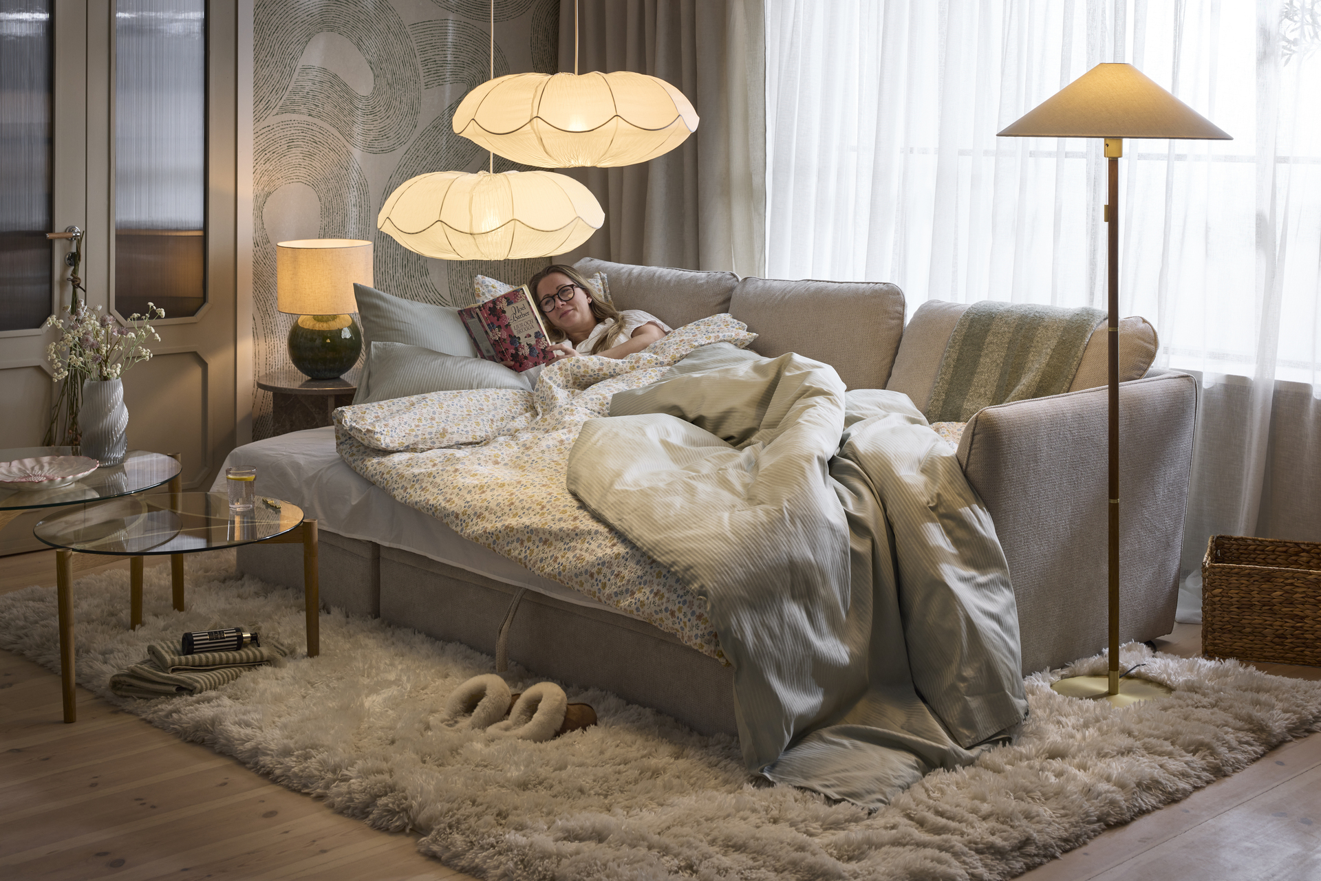 I en bäddsoffa ligger en kvinna nedbäddad under ett täcke och läser en bok