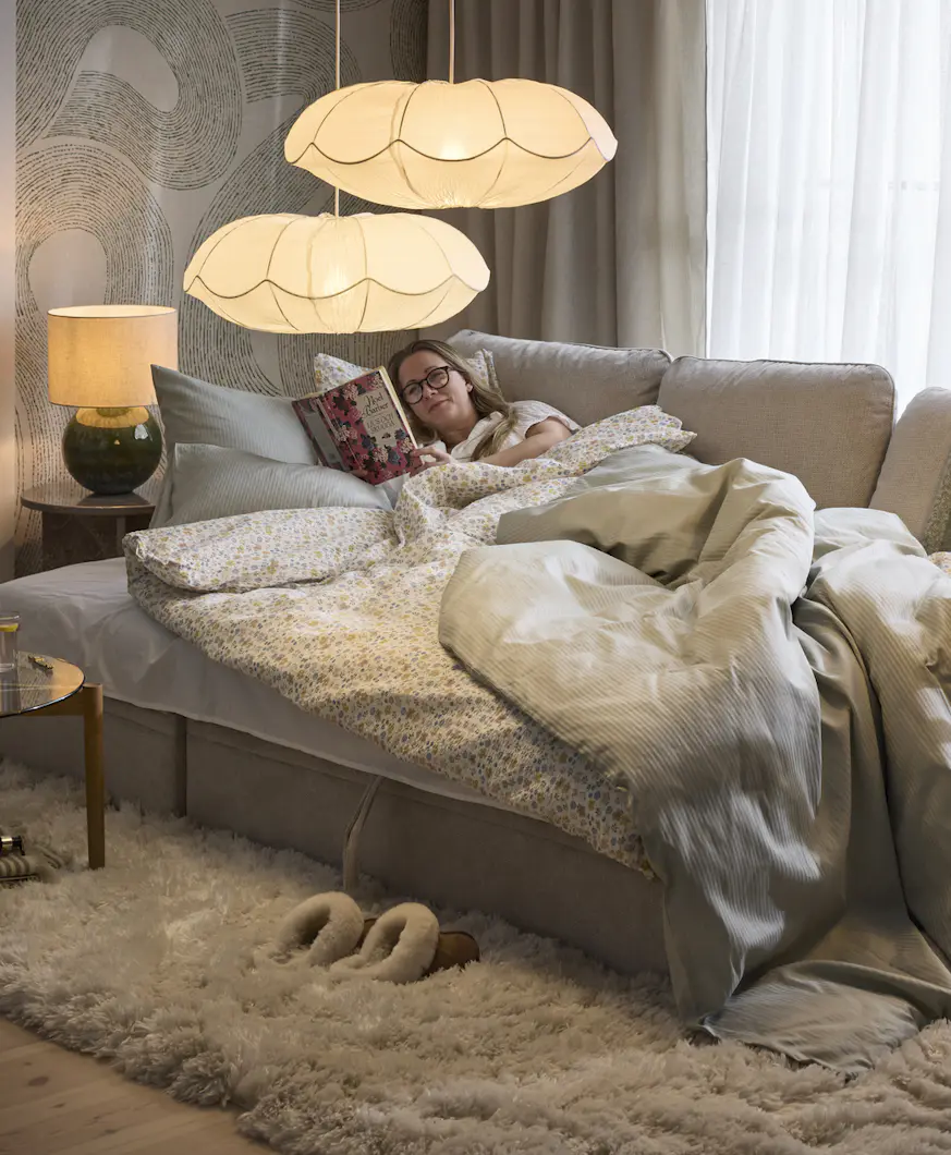 En kvinna ligger nedbäddad i en grå bäddsoffa och läser en bok