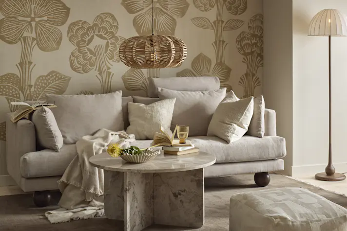 I ett vardagsrum med mönstrad, beige tapet står en beige soffa med klotformade ben