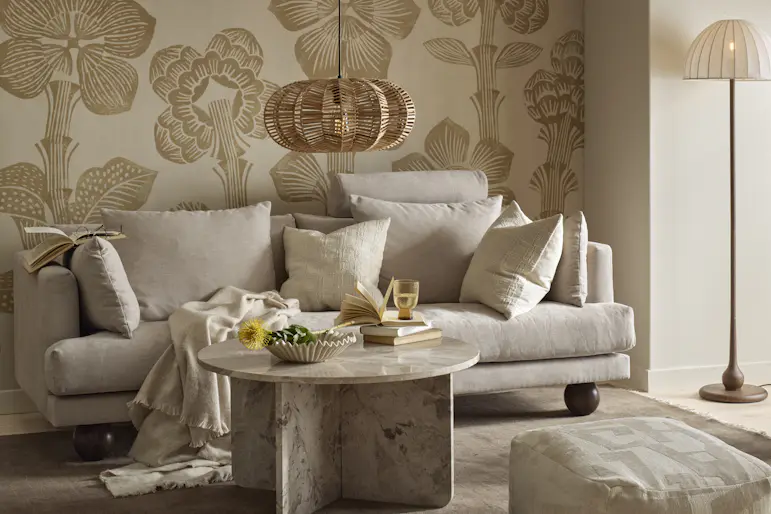I ett vardagsrum med mönstrad, beige tapet står en beige soffa med klotformade ben
