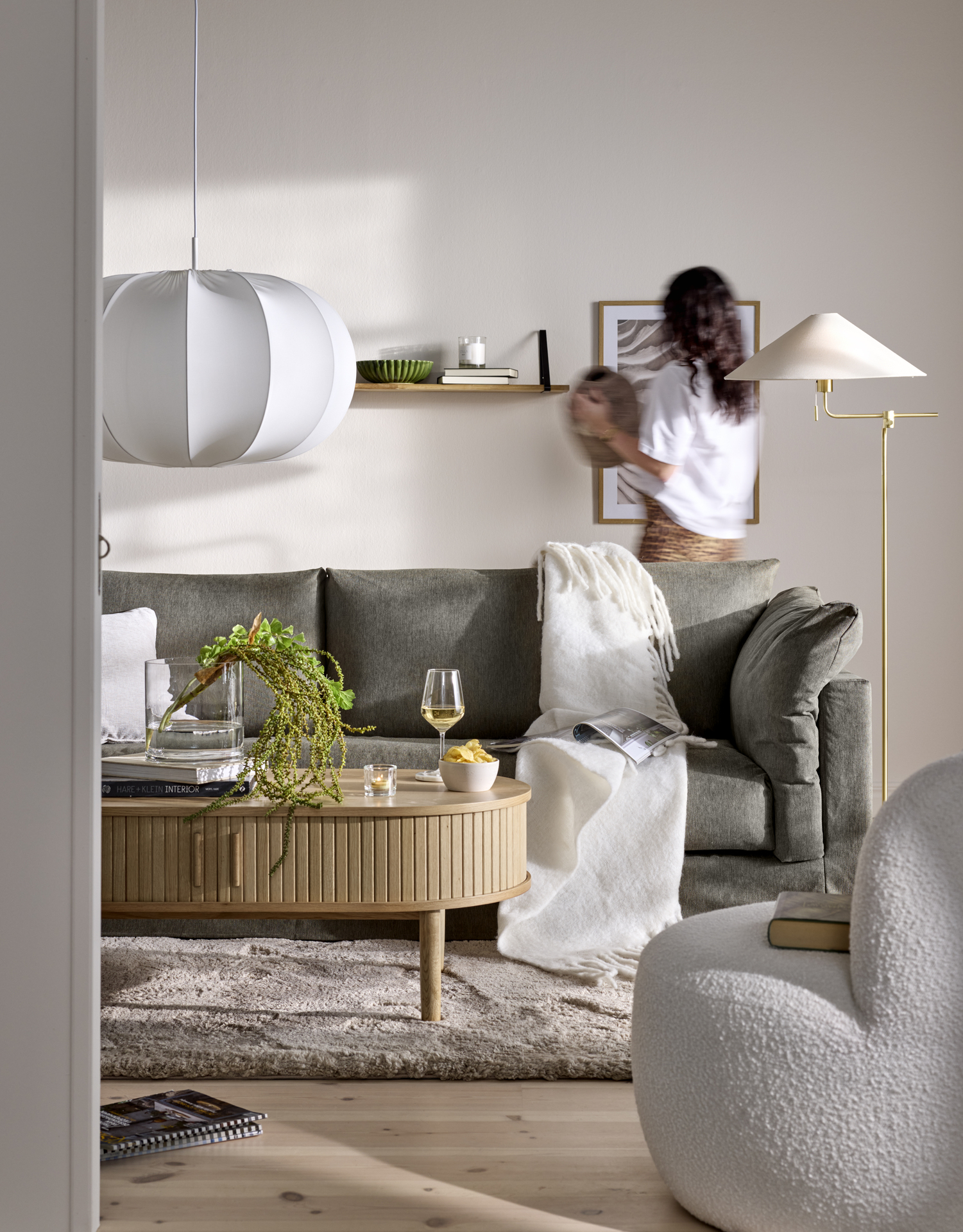En grå Cherrie-soffa i ett beige vardagsrum, bakom soffan står en kvinna