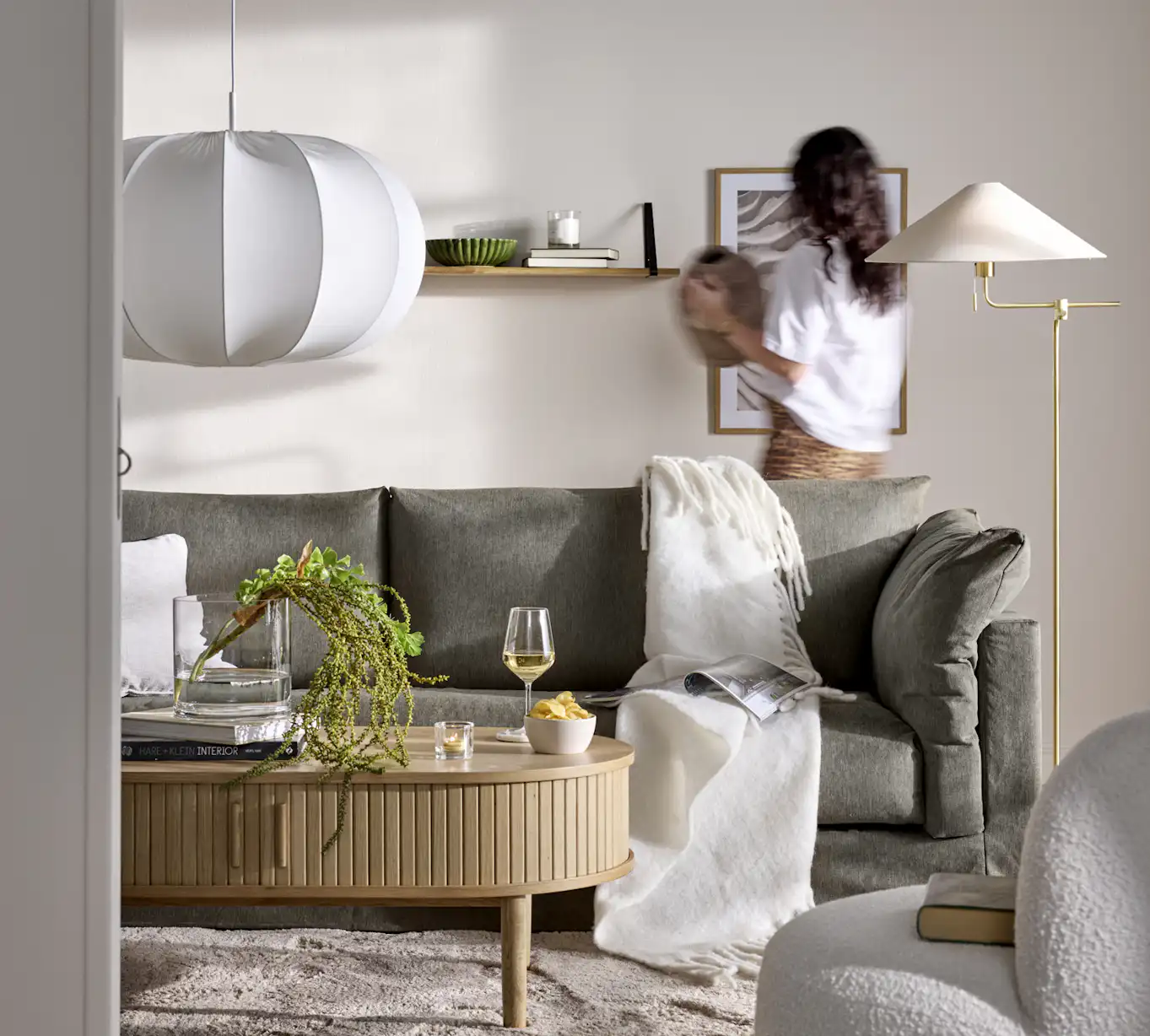 En grå Cherrie-soffa i ett beige vardagsrum, bakom soffan står en kvinna