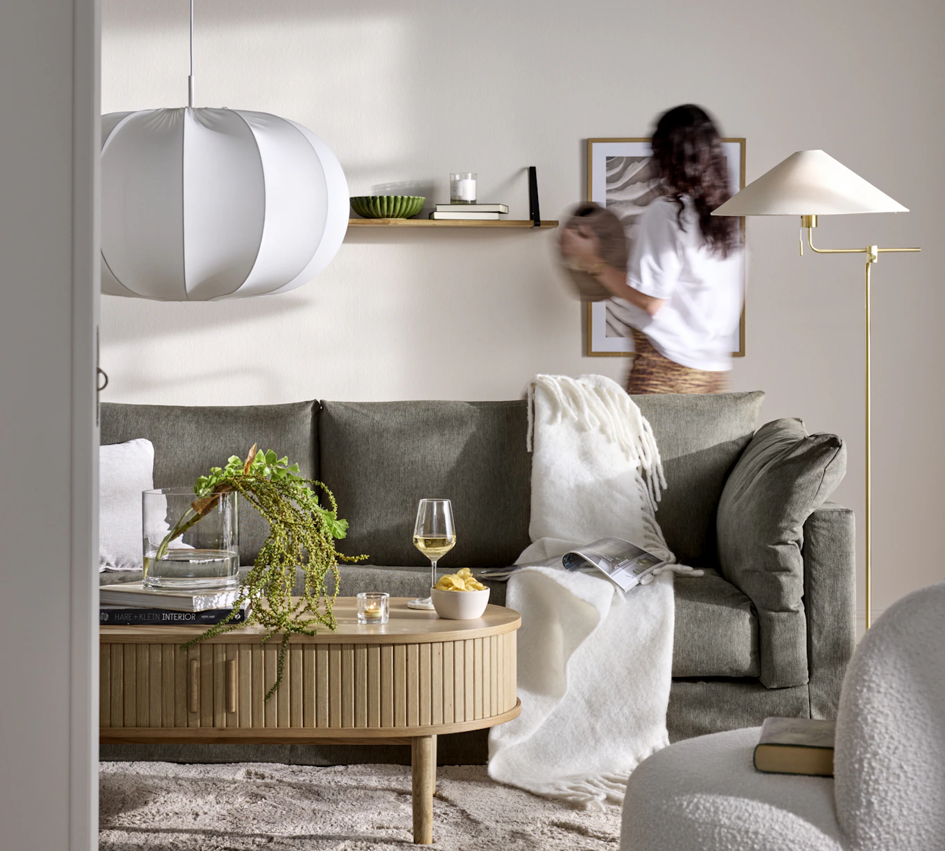 En grå Cherrie-soffa i ett beige vardagsrum, bakom soffan står en kvinna