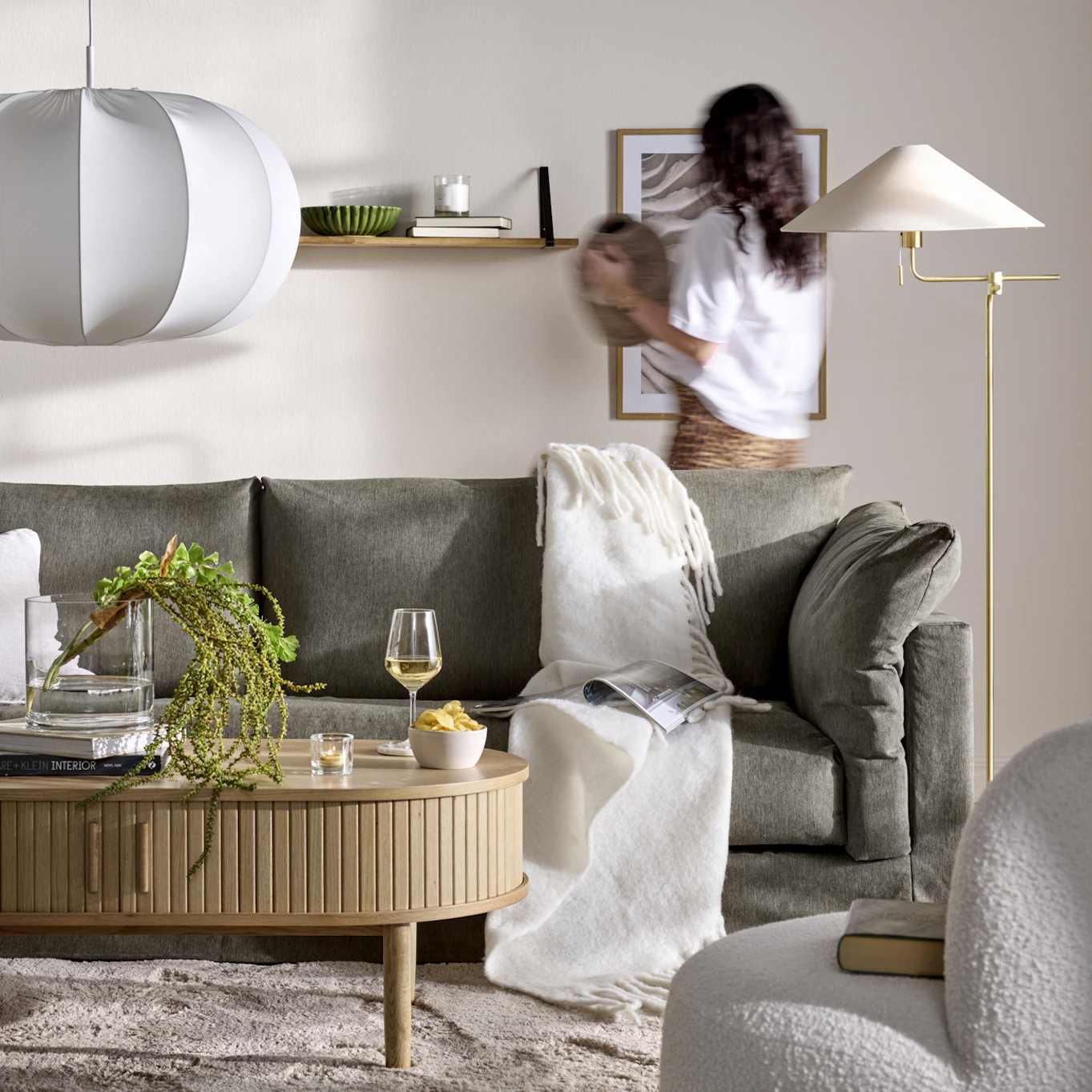 En grå soffa står i ett beige vardagsrum, bakom soffan står en kvinna med en kruka i handen