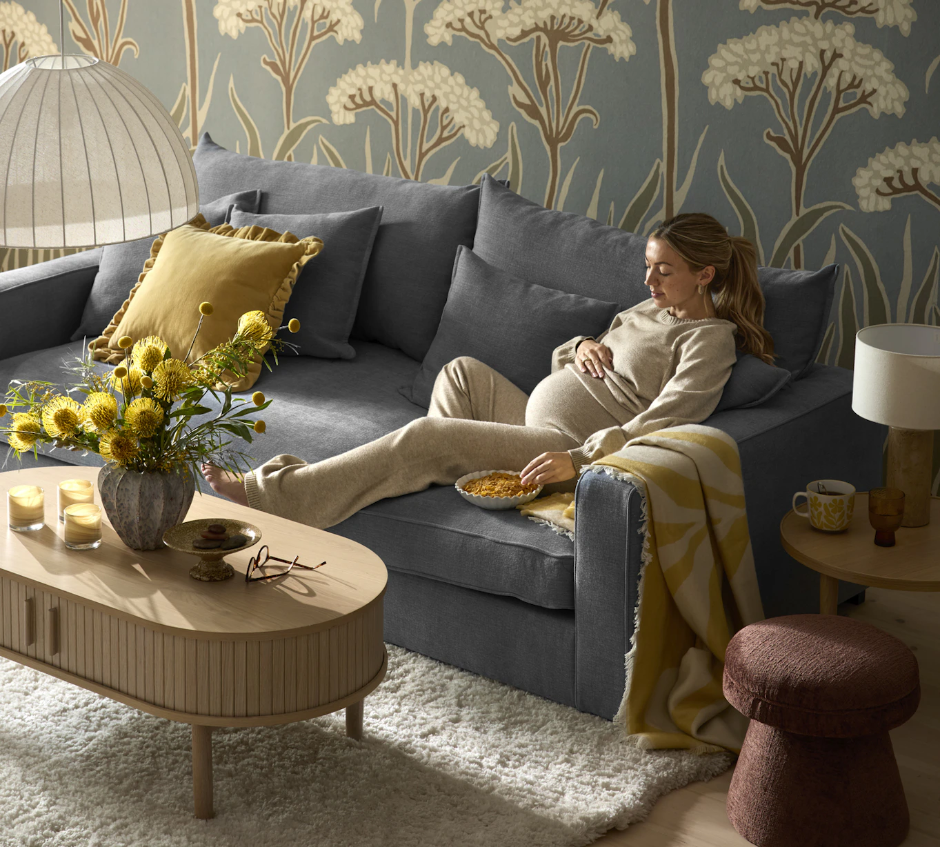 I ett vardagsrum med storblommig tapet sitter en gravid kvinna i en blå, djup soffa.
