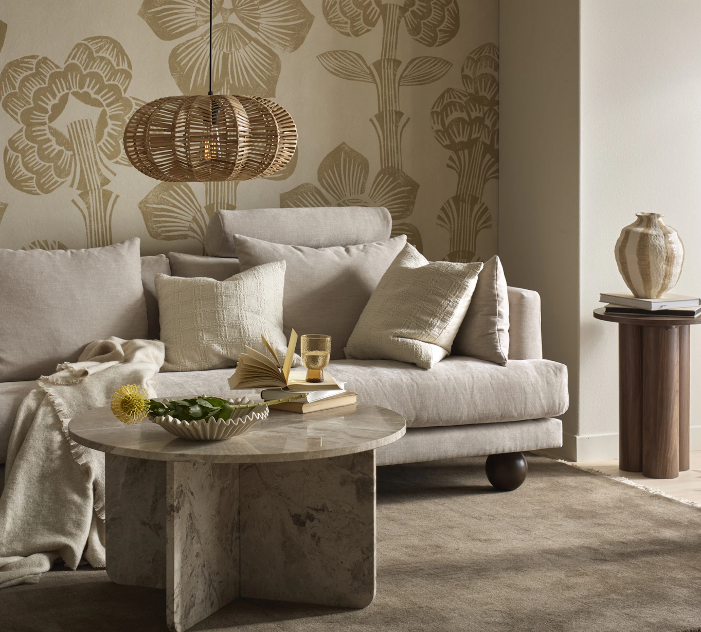 I ett vardagsrum med mönstrad, beige tapet står en beige soffa med klotformade ben