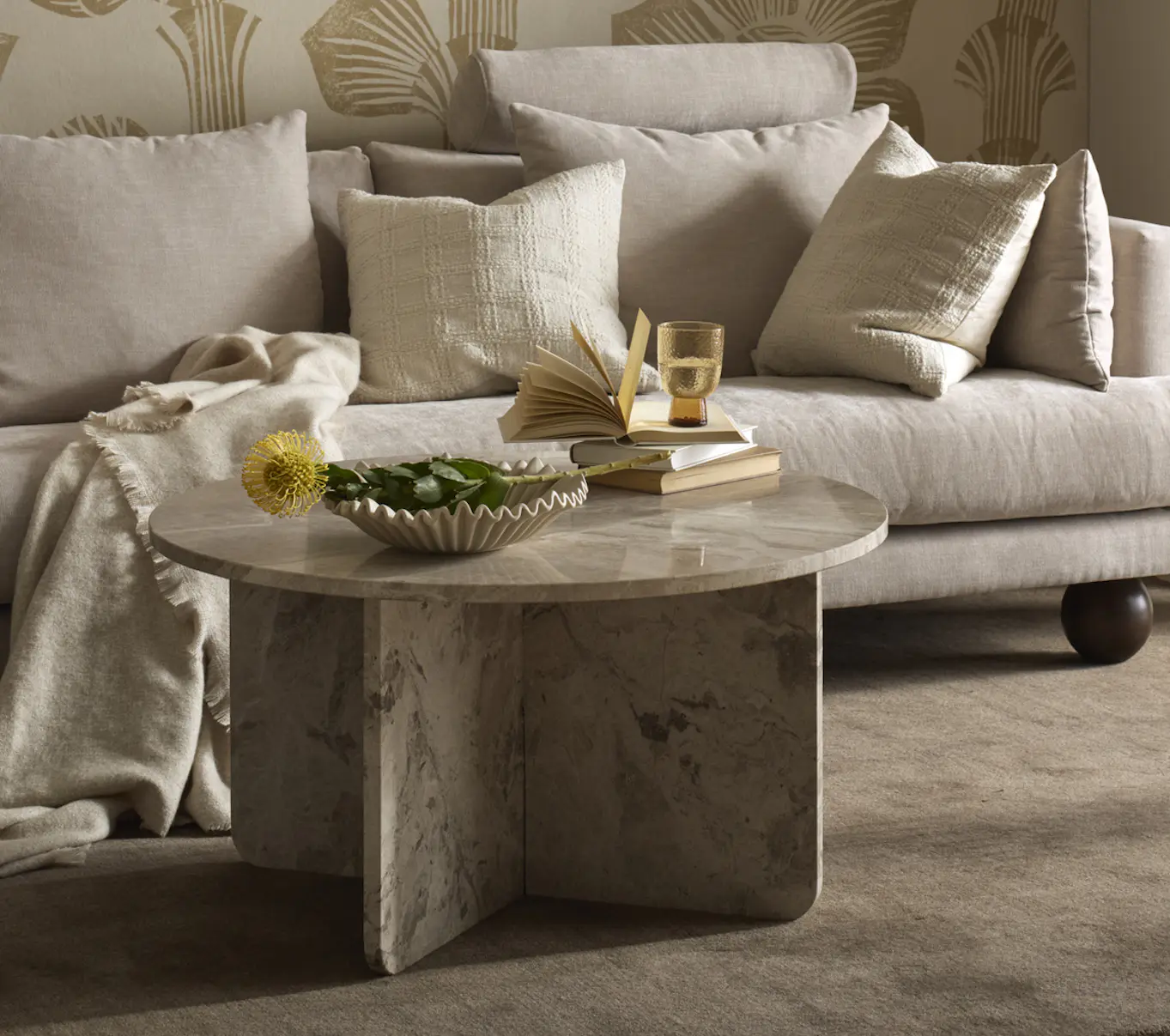 I ett vardagsrum står en beige soffa med klotformade ben bakom ett runt soffbord i beige marmor