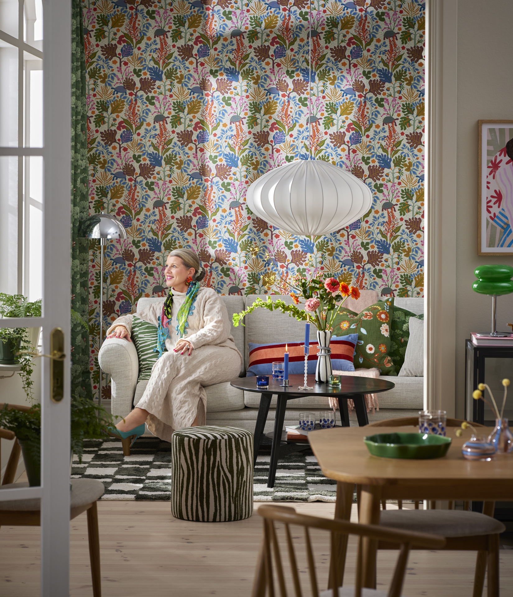 I ett kulört vardagsrum sitter en kvinna i en ljusgrå soff och tittar ut genom ett fönster