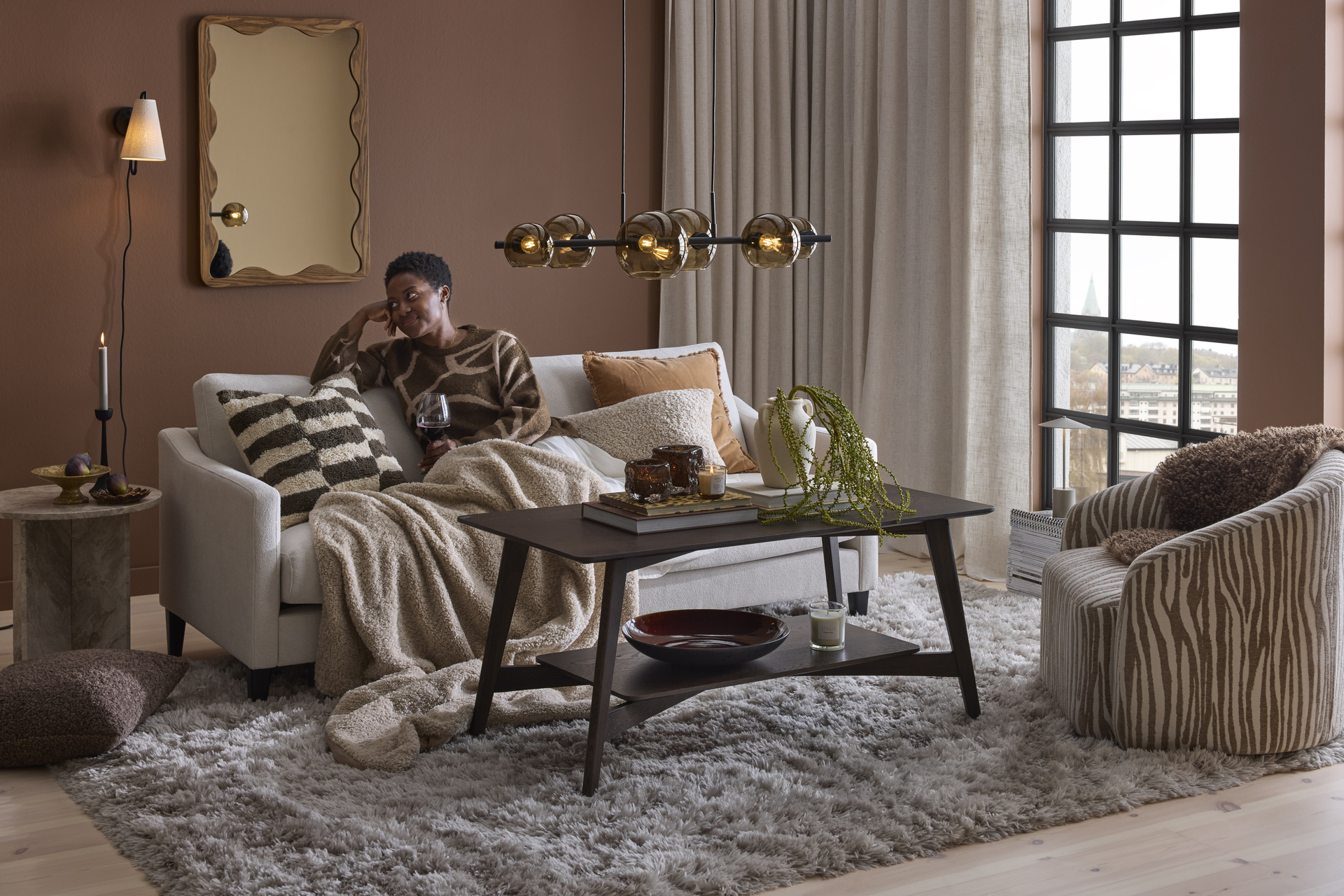 I ett brunt vardagsrum sitter en kvinna under en filt i en ljusgrå soffa 