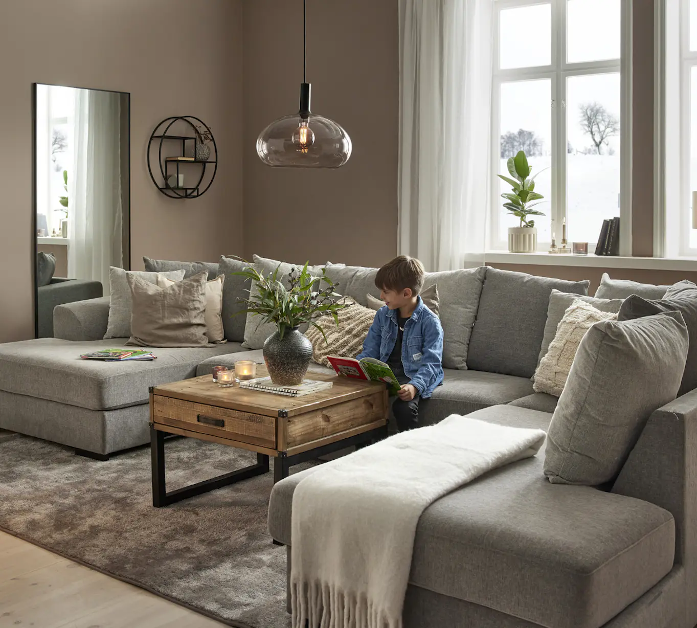I ett vardagsrum står en stor, grå u-soffa, i soffan sitter en pojke och läser