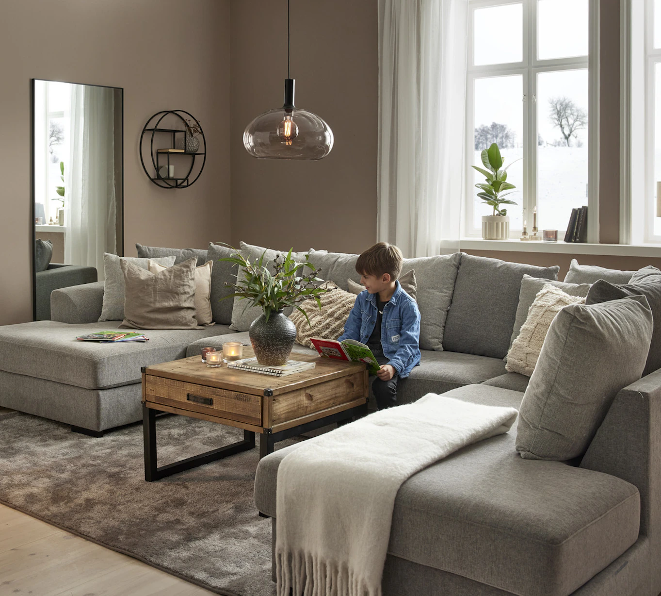 I ett vardagsrum står en stor, grå u-soffa, i soffan sitter en pojke och läser