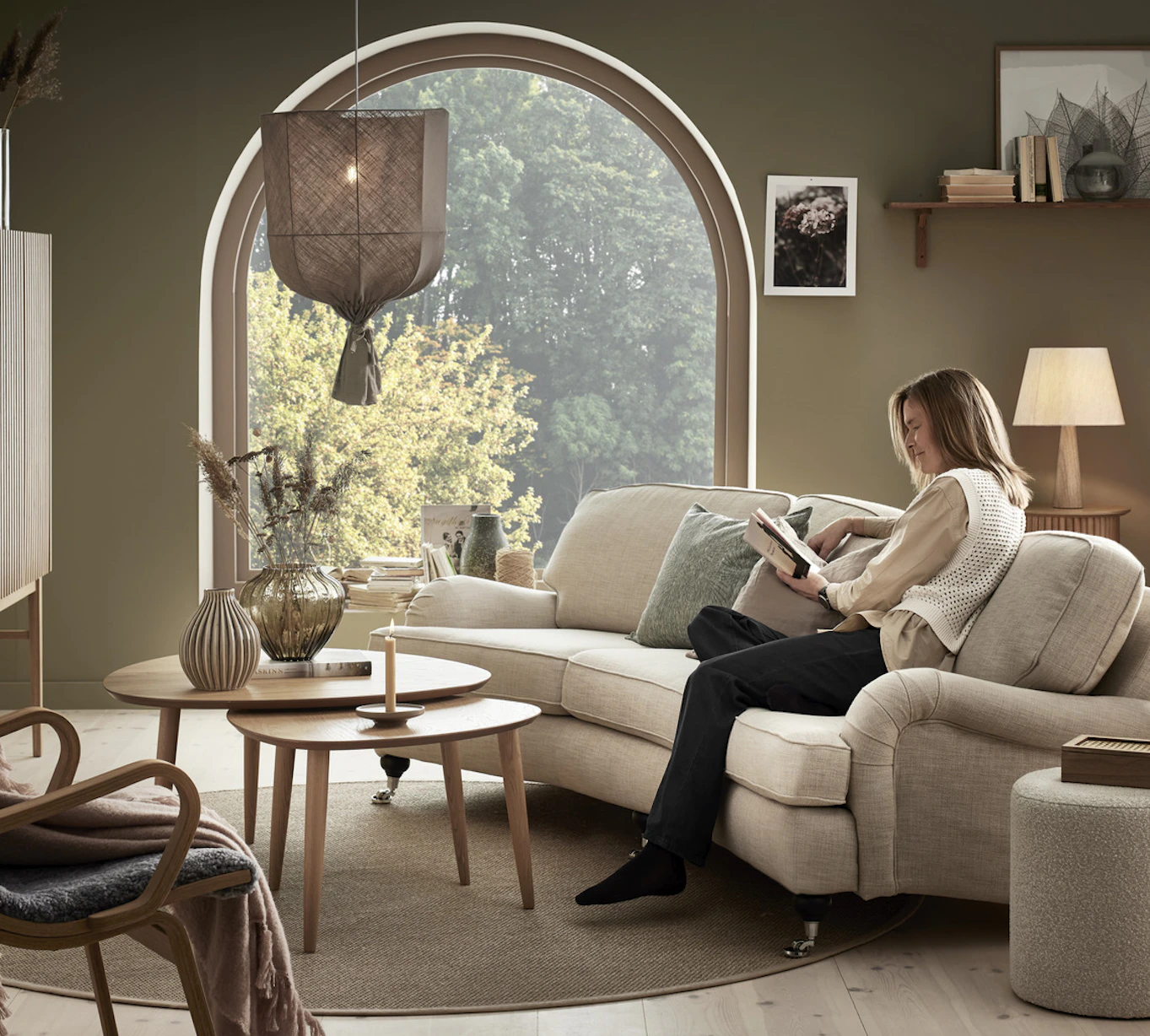I ett grönt vardagsrum sitter en kvinna och läser i en beige Oxford soffa