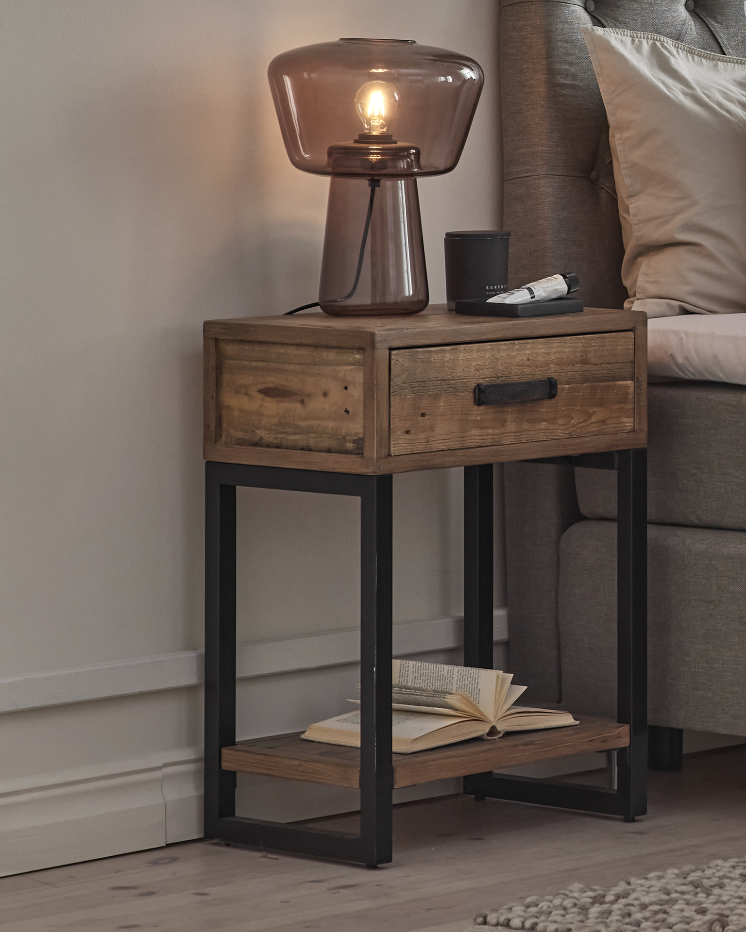 Ett sängbord i rustikt trä med svarta metallben, en låda och en hylla står bredvid en säng . På bordet står en lampa.
