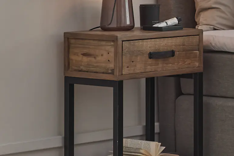 Ett sängbord i rustikt trä med svarta metallben, en låda och en hylla står bredvid en säng . På bordet står en lampa.