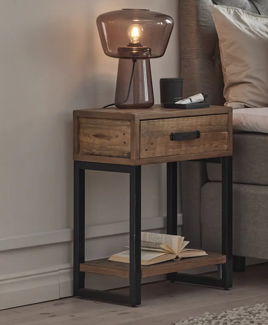 Ett sängbord i rustikt trä med svarta metallben, en låda och en hylla står bredvid en säng . På bordet står en lampa.