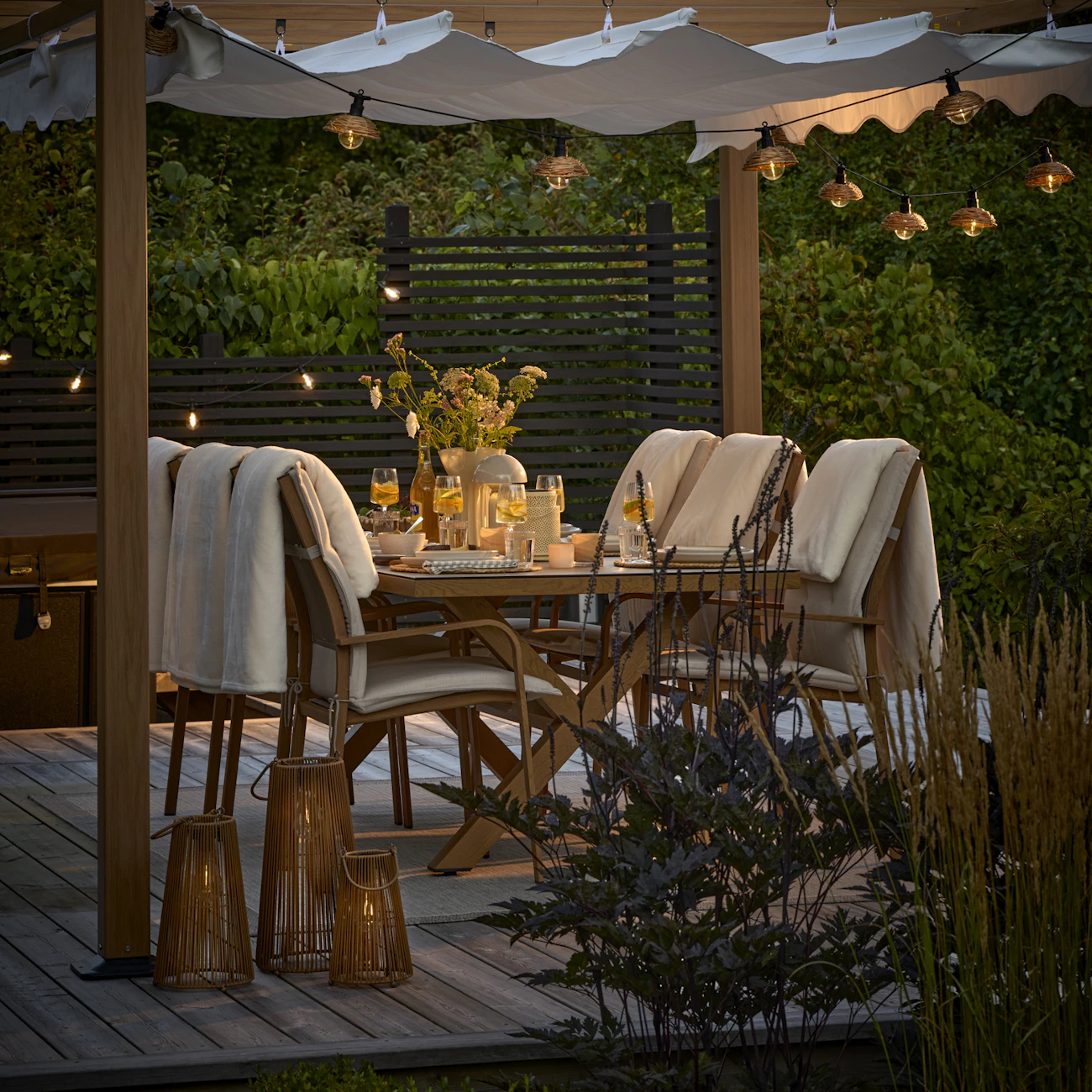 En dukad utemöbelsgrupp står på ett trädäck under en pergola i sommarkvällen.